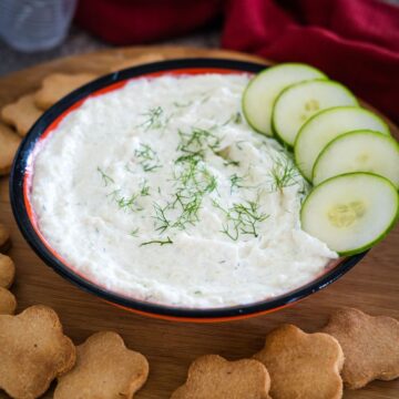 A bowl of creamy dip garnished with dill, accompanied by cucumber slices and flower-shaped crackers on a wooden board.