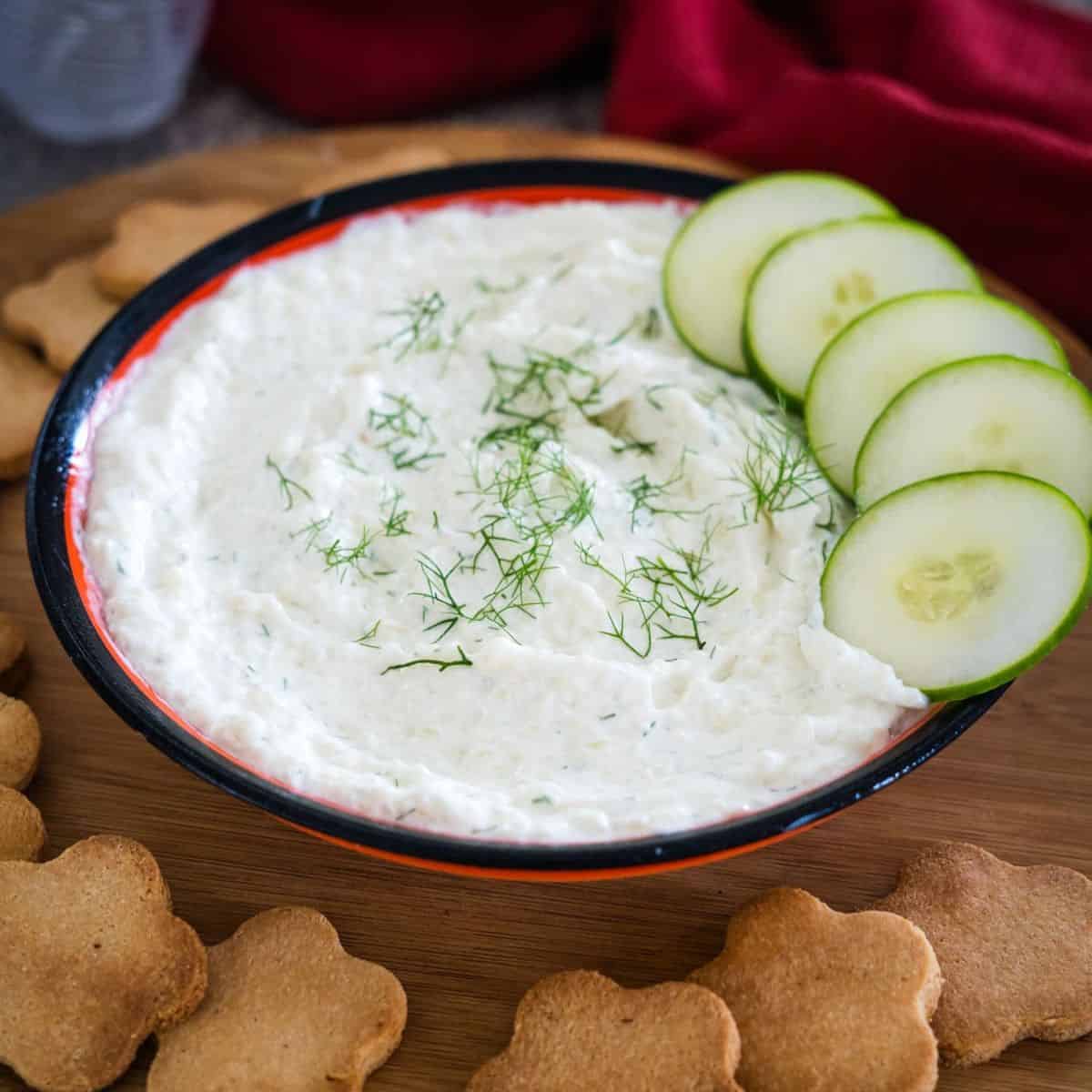 A bowl of creamy dip garnished with dill, accompanied by cucumber slices and flower-shaped crackers on a wooden board.