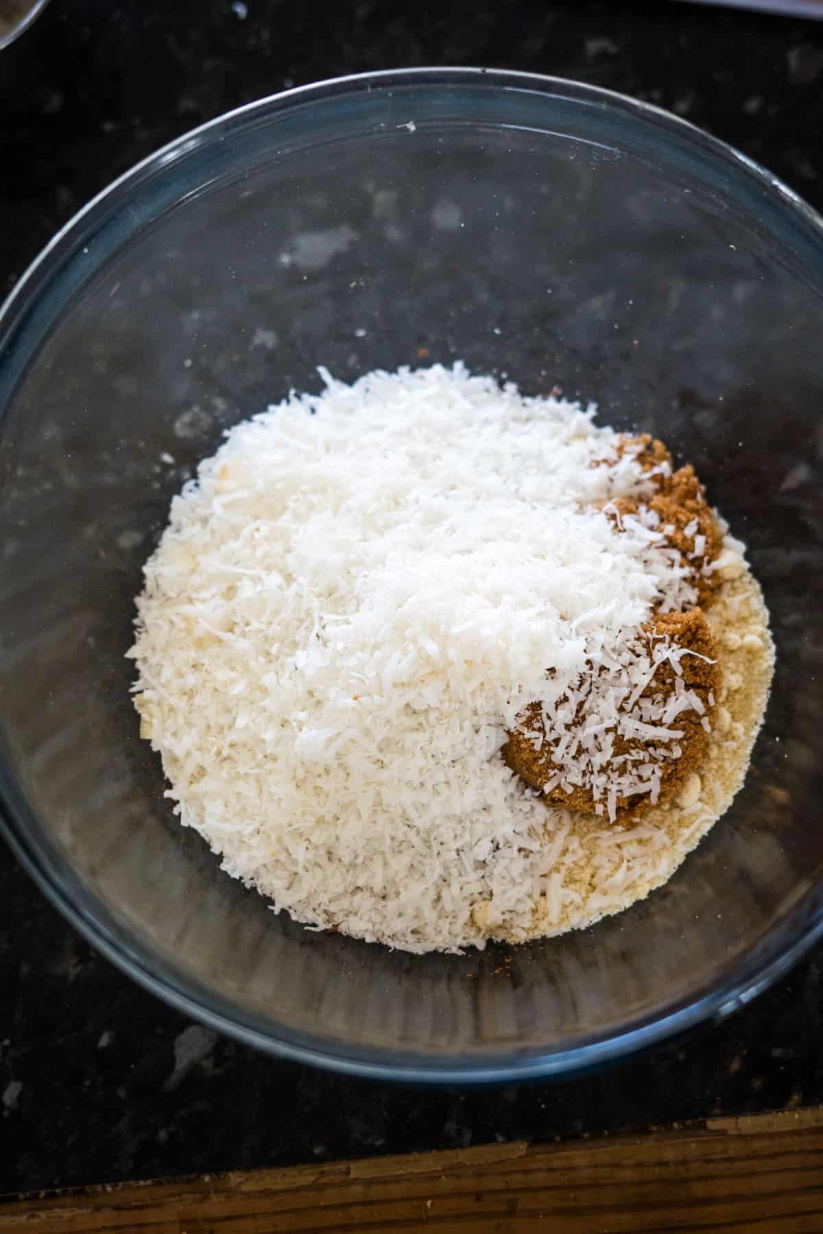 A clear glass bowl containing a mixture of shredded coconut, brown sugar, and powdered ingredients on a dark countertop.