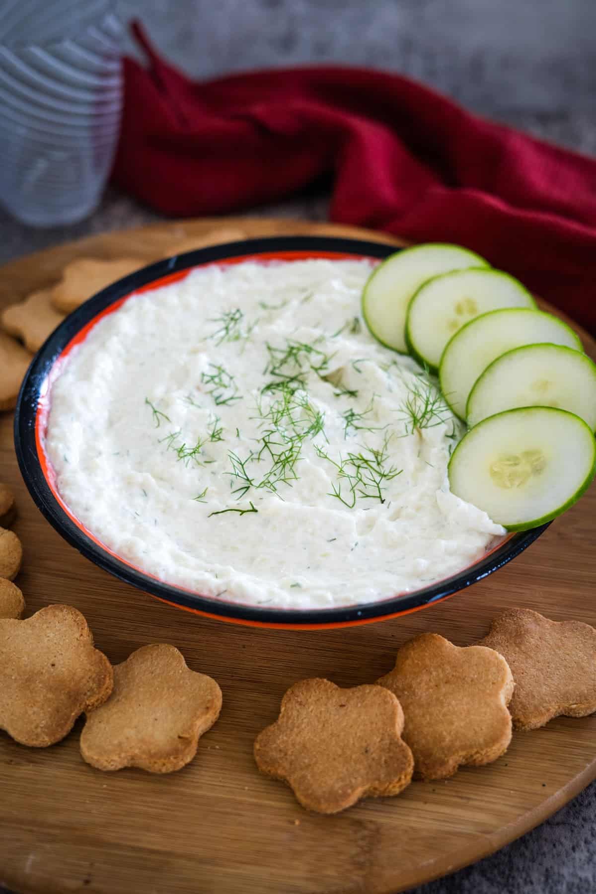 A black bowl filled with cucumber cream cheese dip garnished with dill and cucumber slices, placed on a wooden board surrounded by flower-shaped crackers. A red cloth is in the background.