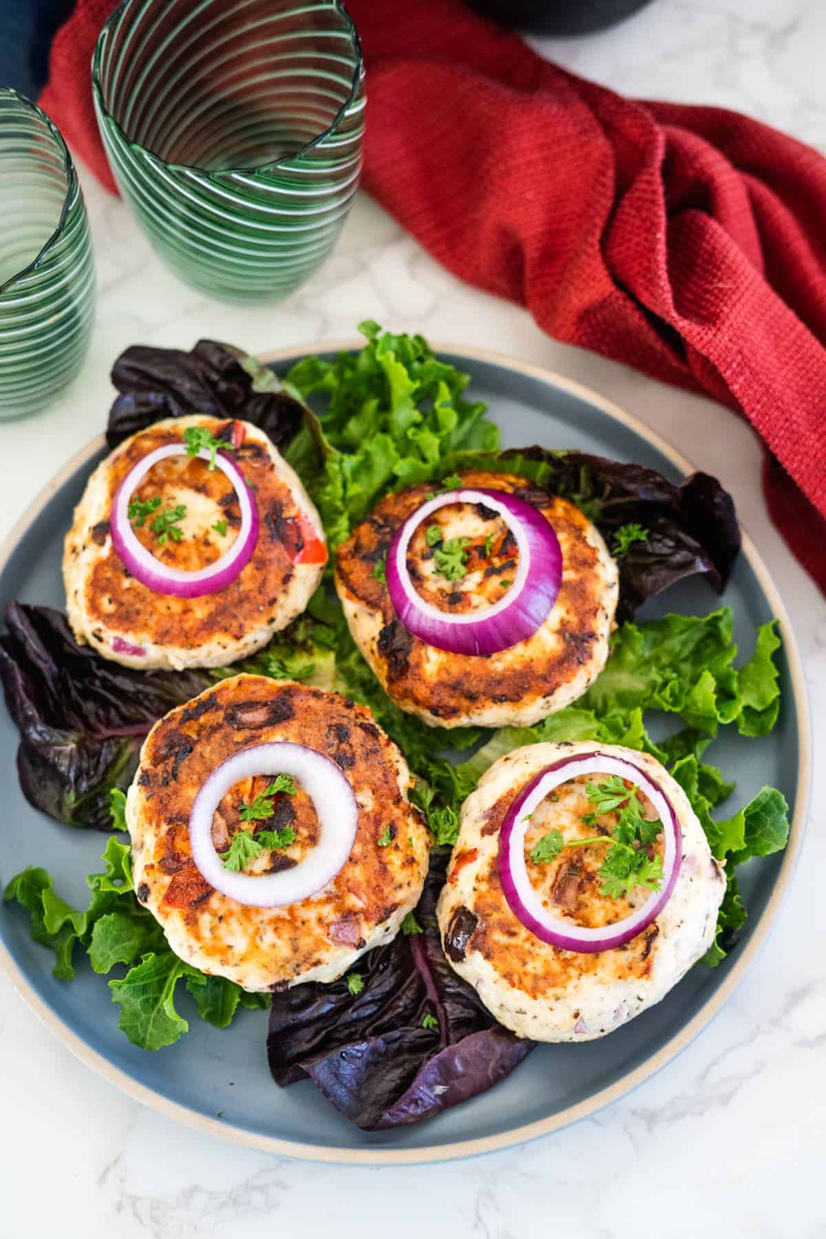 A plate with four grilled chicken patties garnished with red onion rings and fresh parsley, served on a bed of mixed greens. Two striped glass tumblers and a red cloth napkin are in the background.