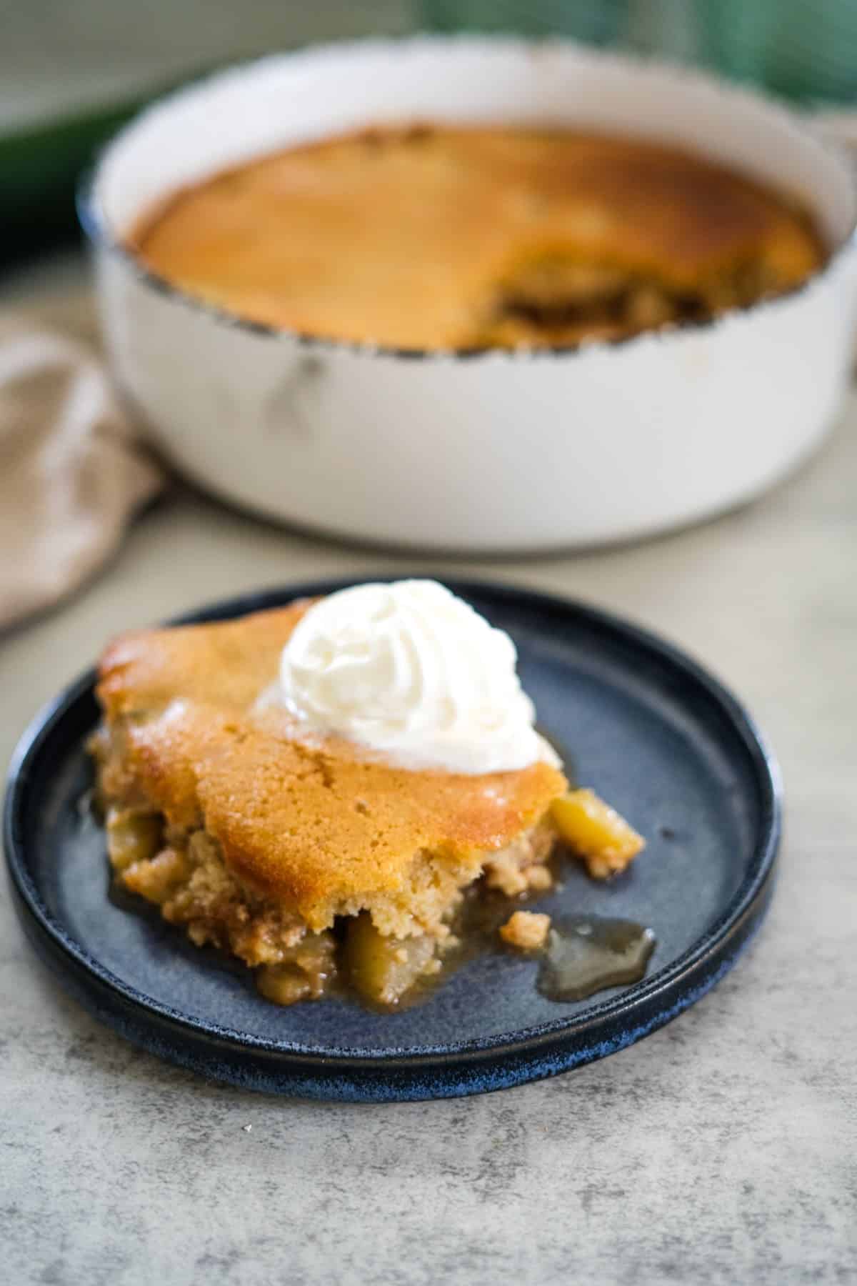 A slice of zucchini cobbler topped with whipped cream sits on a blue plate, with the remaining cobbler in a white dish in the background.