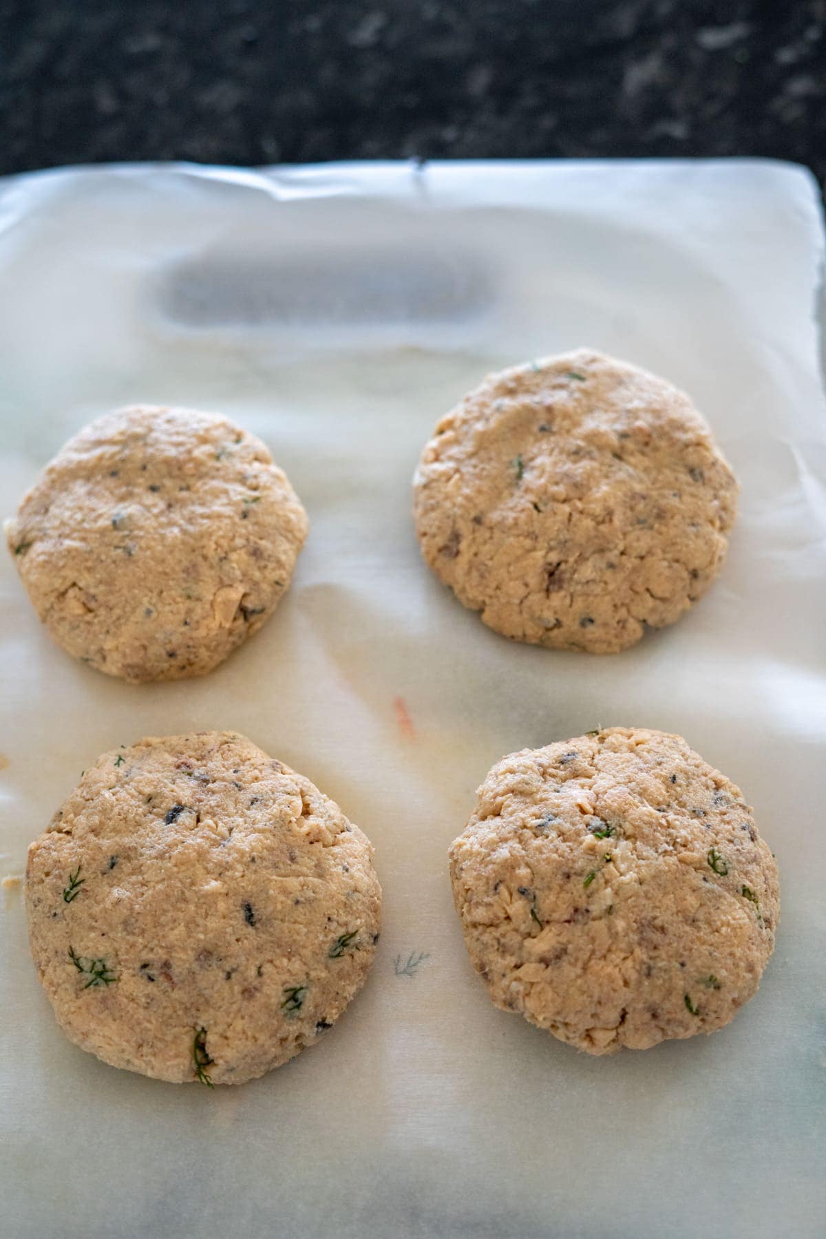 Four uncooked fish patties on a sheet of parchment paper with a dark background.