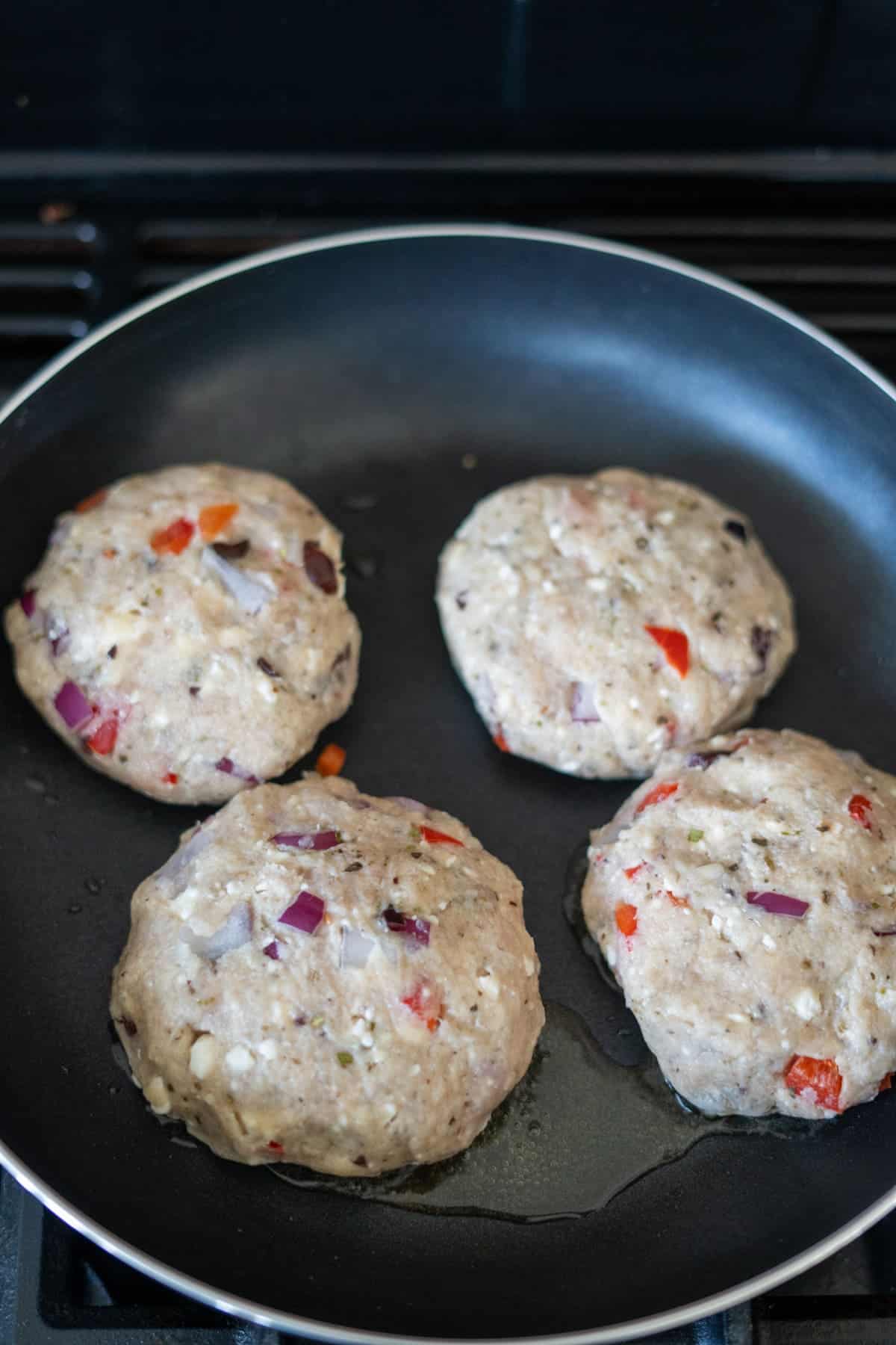 Four uncooked Greek chicken burgers with chopped vegetables are sizzling in a black frying pan.