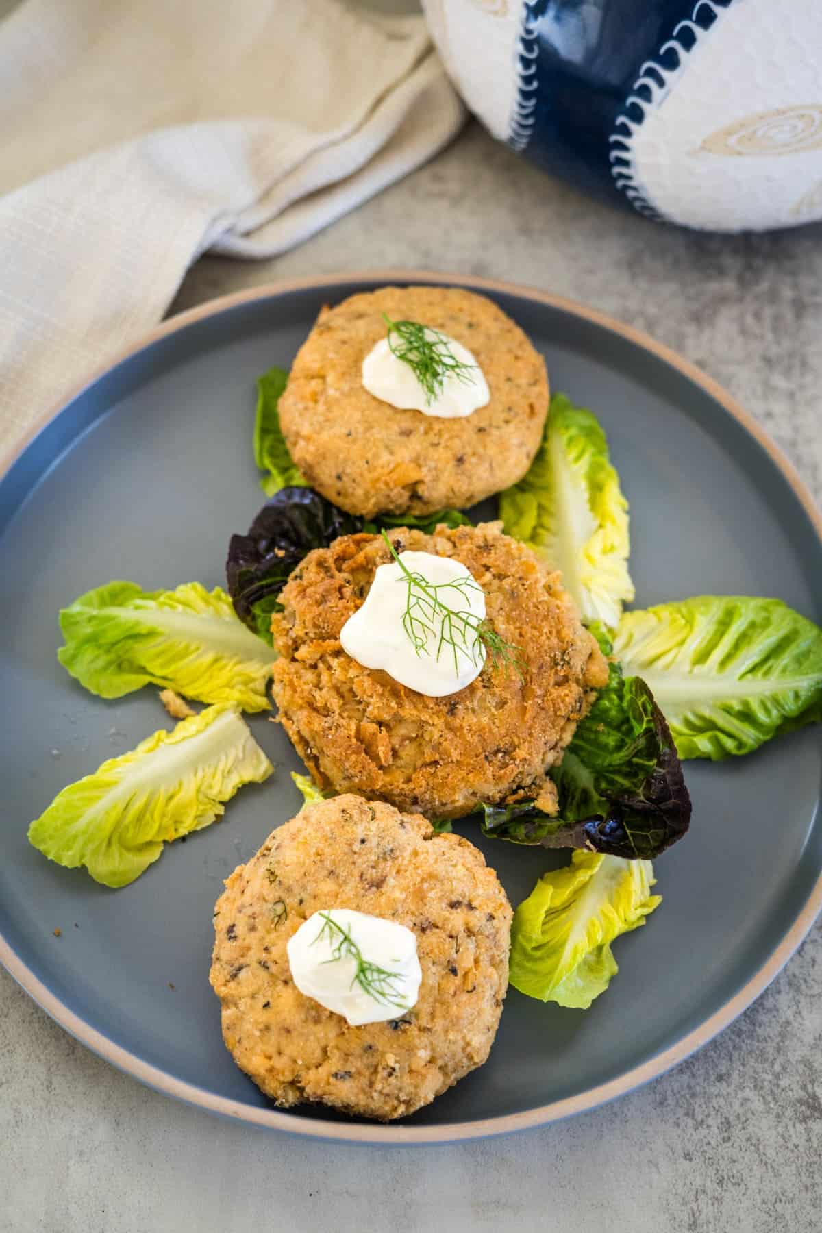 Three crab cakes topped with a dollop of sauce and a dill sprig are placed on a grey plate with a bed of lettuce on a light-colored table.