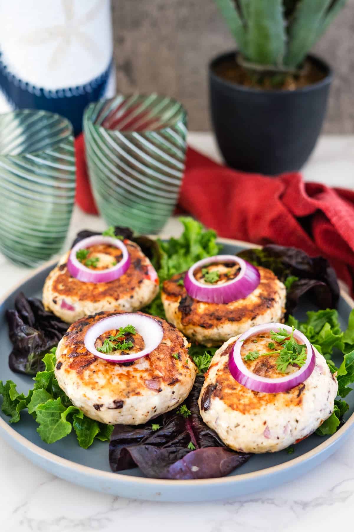 Four Greek chicken burgers topped with red onion rings and herbs are served on a bed of leafy greens on a green plate. Two striped glasses, a cactus in a black pot, and a red cloth are in the background.