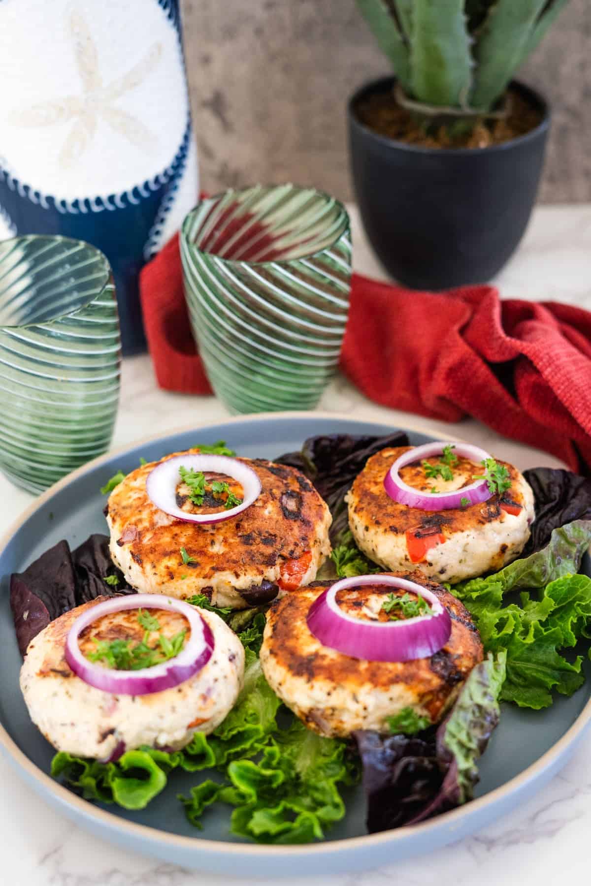 A plate with four Greek chicken burgers topped with red onion slices and cilantro, served on a bed of lettuce. Two green glasses and a potted cactus are in the background.