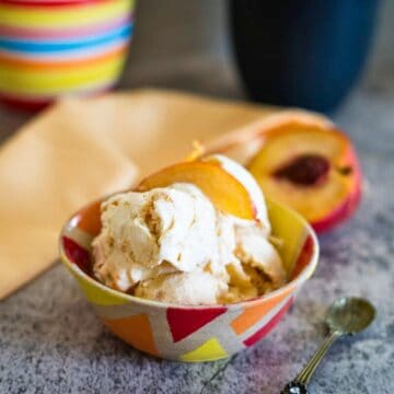 A bowl of ice cream topped with peach slices sits on a table, with a spoon beside it and a colorful cup and napkin in the background.