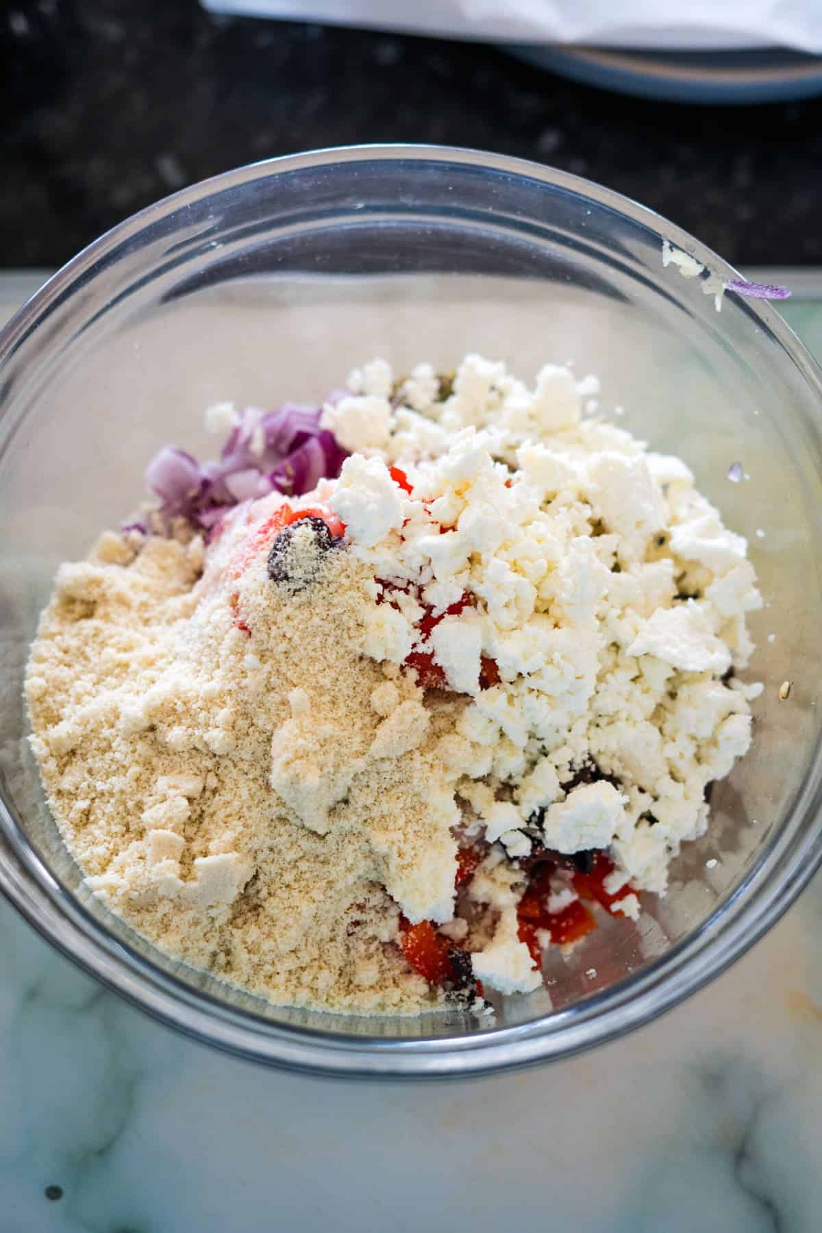 A glass bowl containing a mixture of crumbled feta cheese, breadcrumbs, chopped red onions, and red bell peppers awaits to be transformed into delicious Greek chicken burgers.