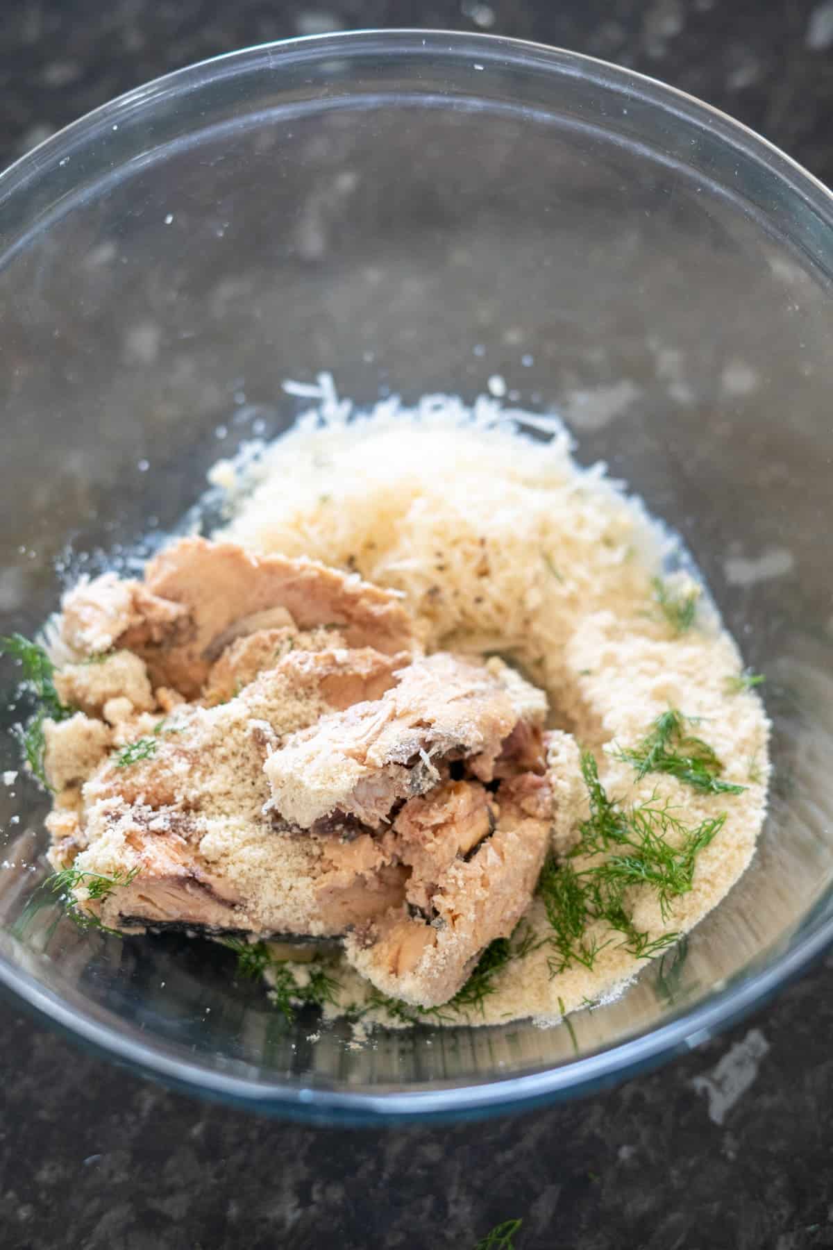 A glass bowl containing shredded cheese, breadcrumbs, tuna, and dill on a dark speckled countertop.