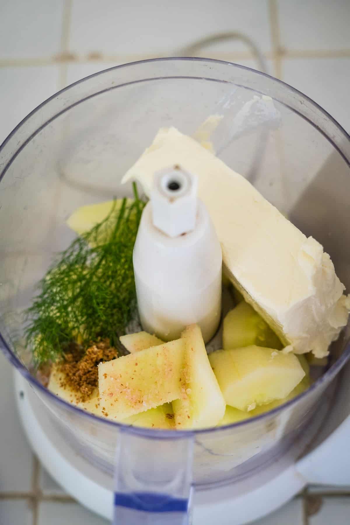 Close-up of a food processor containing a stick of butter, fennel fronds, sliced apples, spices, and a hint of cucumber cream cheese dip, all ready to be blended together.