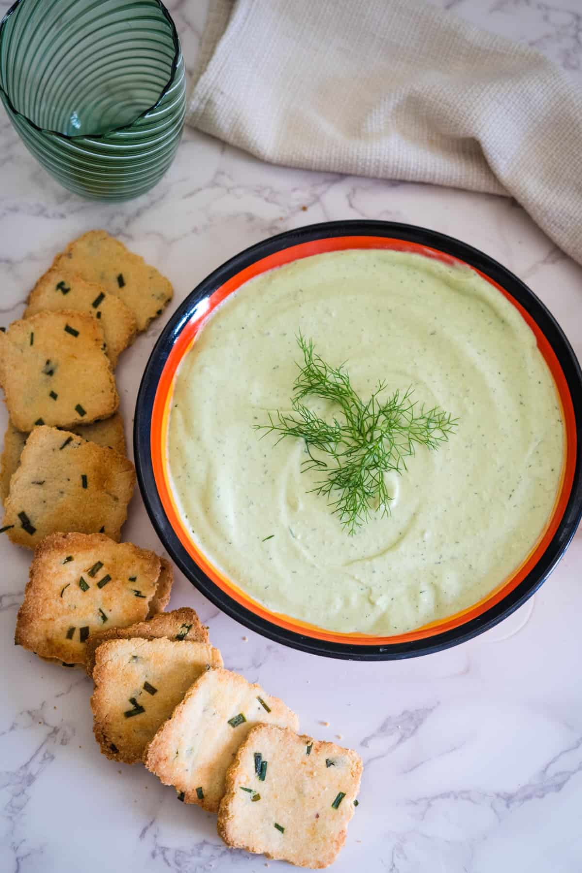 A bowl of creamy green dip garnished with dill sits on a marble surface, accompanied by several rectangular slices of herb-topped bread on the left side. A green glass and a cloth are in the background.