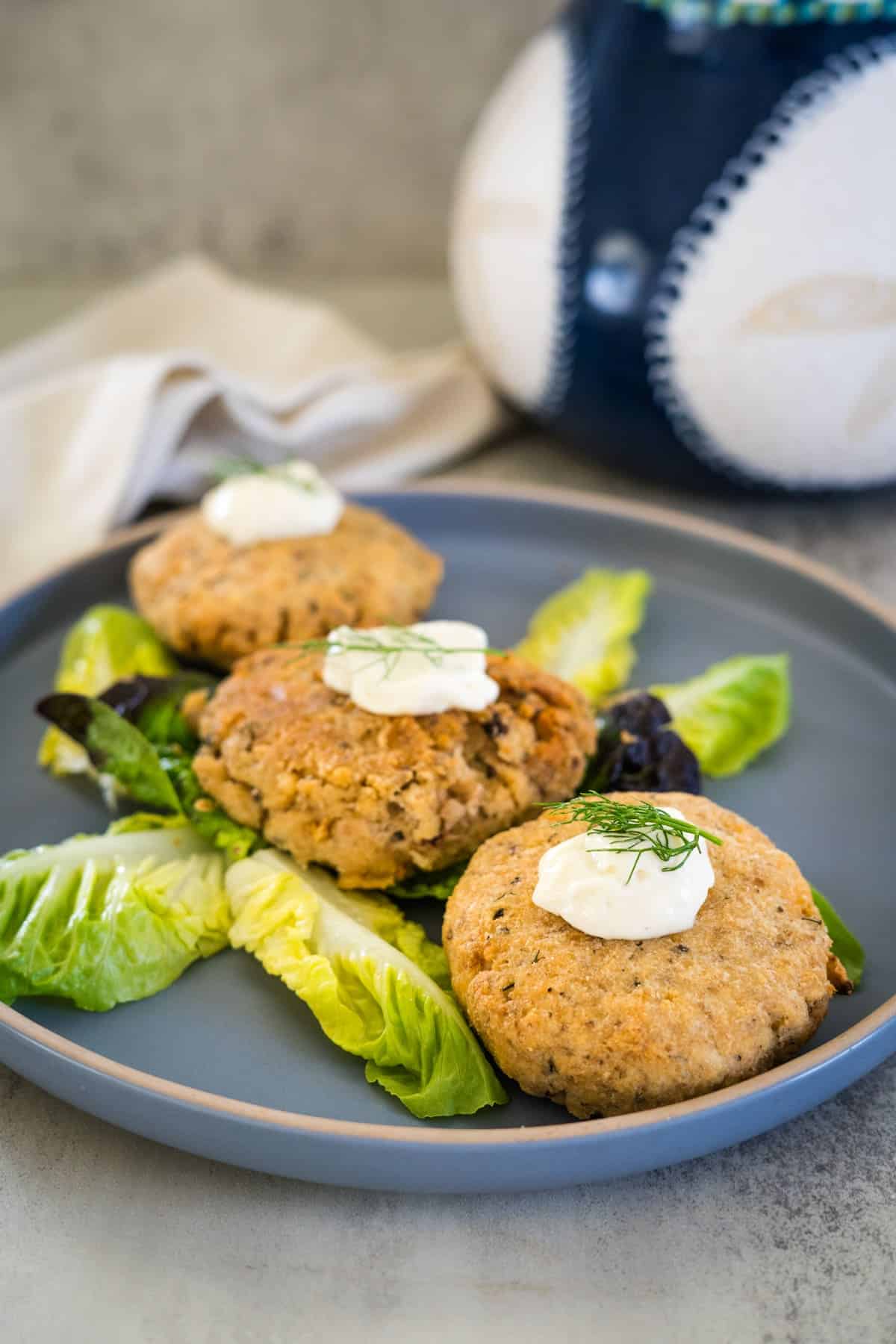 A plate with three fish cakes topped with dollops of white sauce, garnished with fresh dill, and served on a bed of leafy greens. A napkin and ceramic pottery are visible in the background.