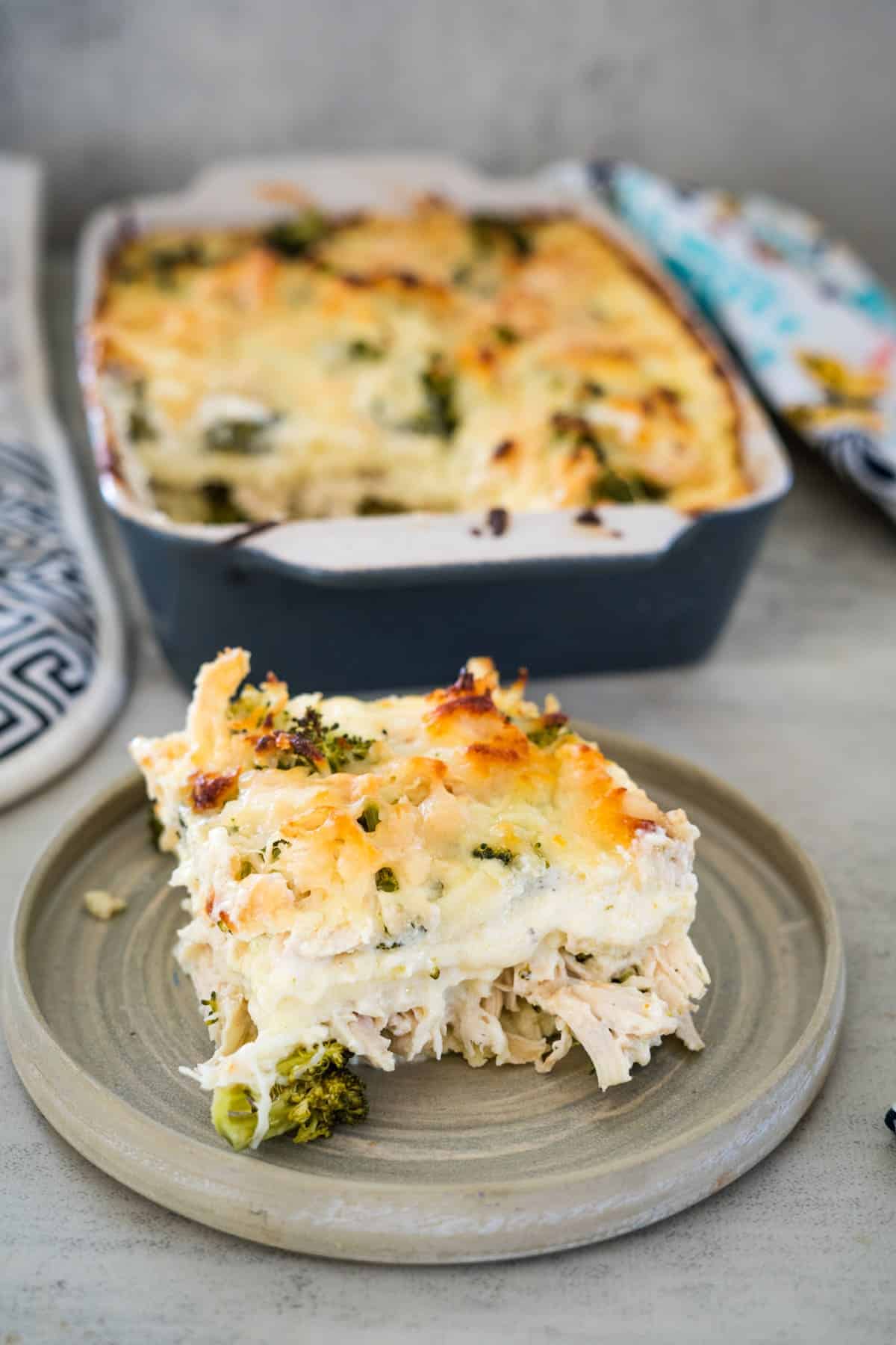 A slice of creamy chicken broccoli lasagna casserole on a plate, with the rest of the dish in a baking pan in the background.