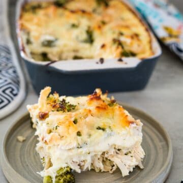 A slice of creamy chicken and broccoli casserole topped with melted cheese is served on a plate, featuring delightful layers of keto lasagna noodles. The remaining casserole rests invitingly in a baking dish in the background.