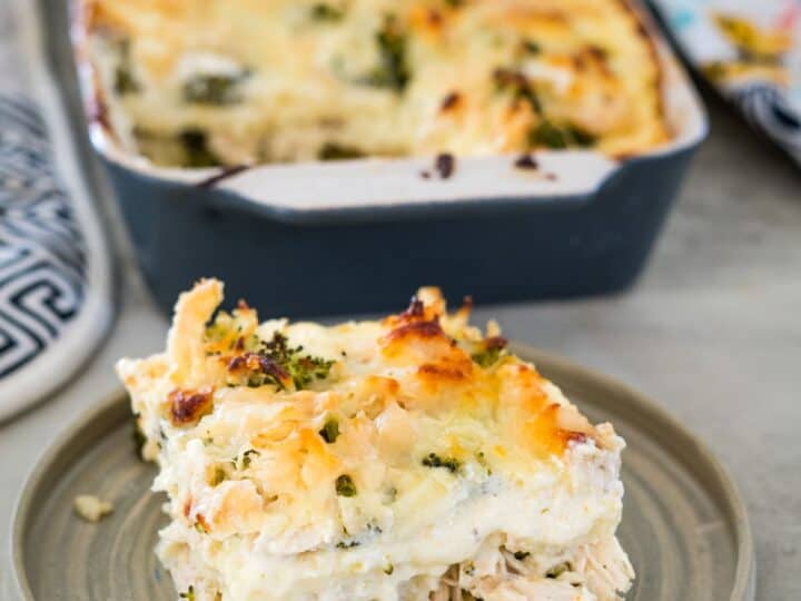 A slice of creamy chicken and broccoli casserole topped with melted cheese is served on a plate, featuring delightful layers of keto lasagna noodles. The remaining casserole rests invitingly in a baking dish in the background.