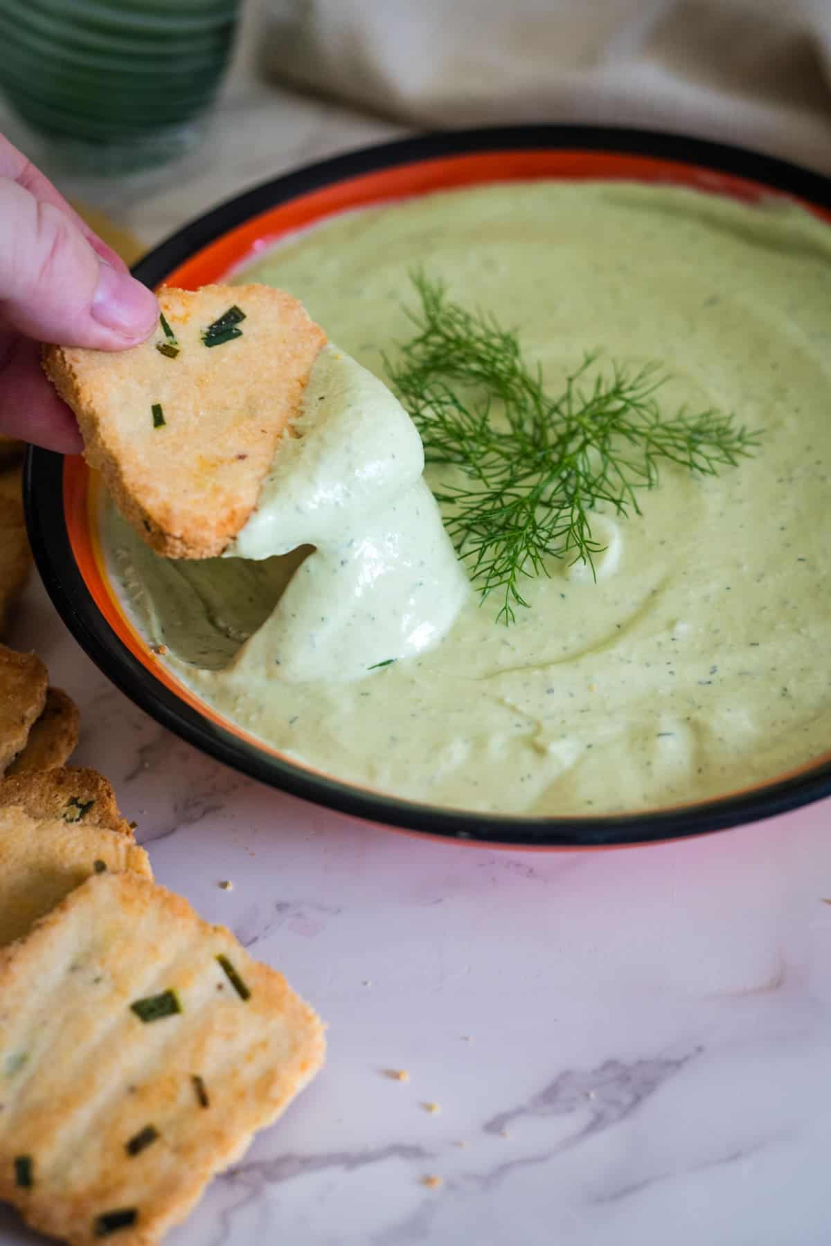 A hand dips a piece of cracker into a bowl of green dip, garnished with a sprig of dill, on a marble surface. Other crackers are placed around the bowl.