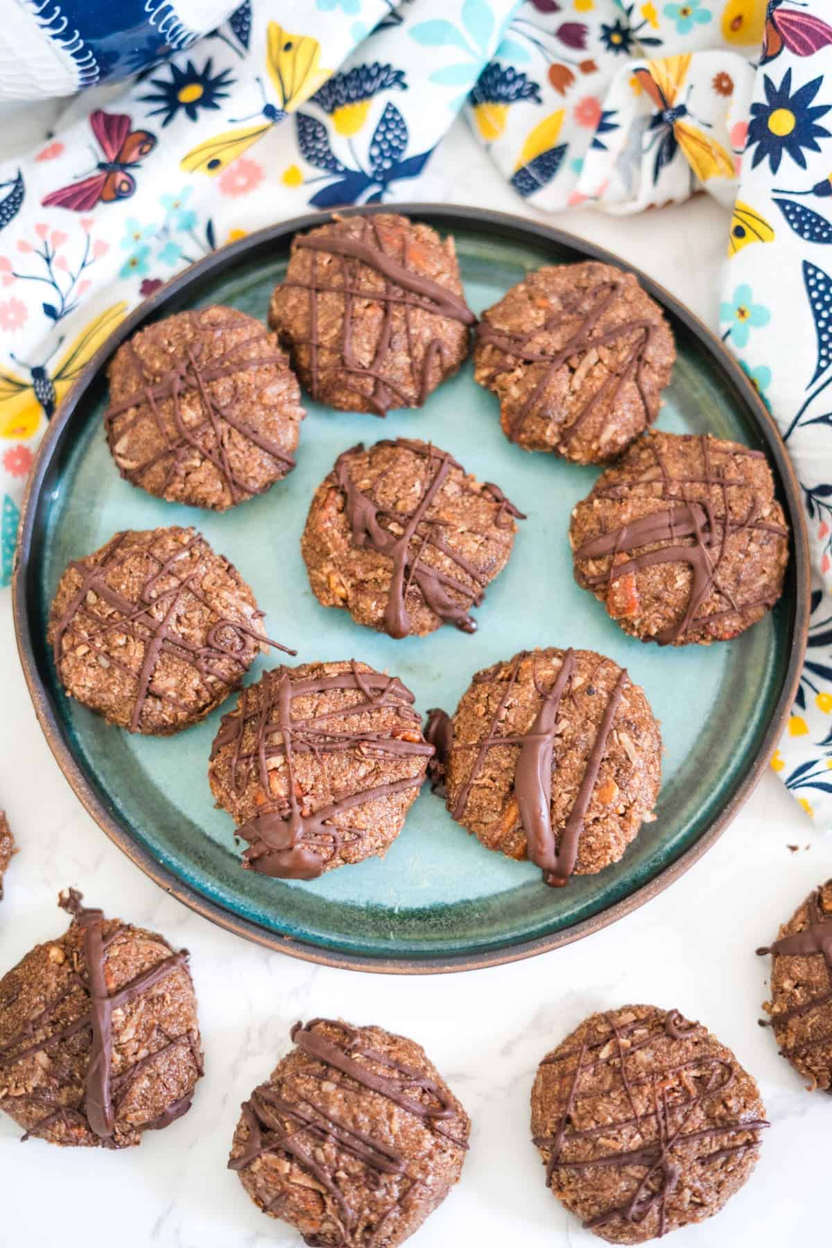 A turquoise plate holds ten chocolate-drizzled oatmeal cookies, surrounded by additional cookies on a white surface. The background features a colorful floral-patterned cloth.