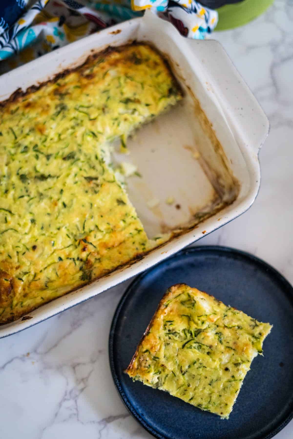 A rectangular dish of baked zucchini casserole with a piece cut out and placed on a black plate beside it. The casserole is light yellow-green with a golden-brown top.