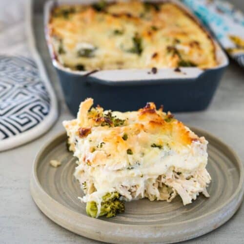 A slice of creamy, baked pasta dish with chicken and broccoli on a round plate. The remainder of the dish is in the background in a rectangular baking pan.