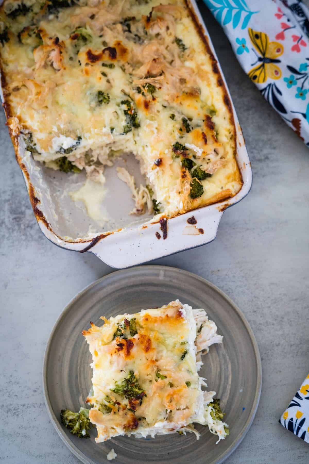 A casserole dish of chicken broccoli lasagna bake with a portion removed and placed on a grey plate. A floral-patterned cloth is partially visible in the background.