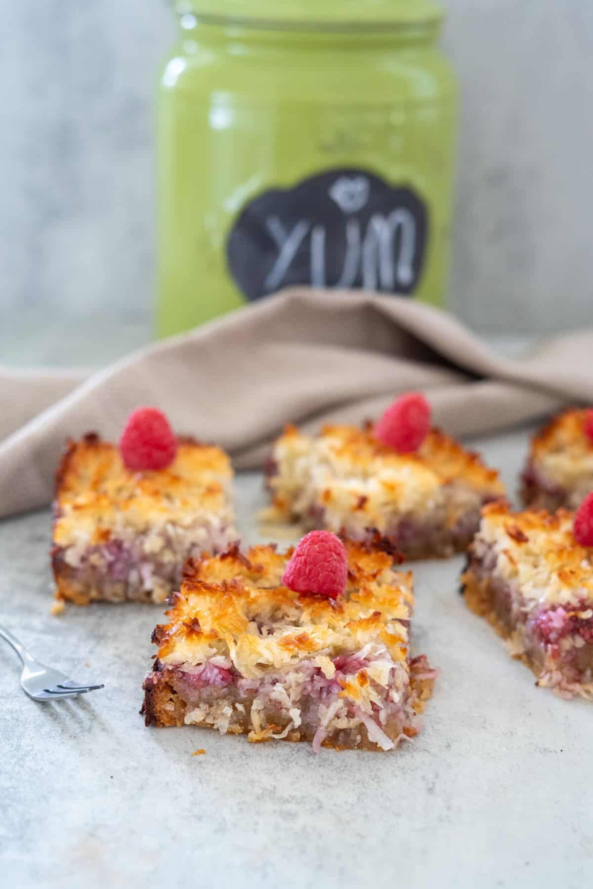 Five square dessert bars topped with raspberries in front of a green jar labeled "Yum" on a light-colored surface. A beige napkin is draped nearby, with a small fork placed beside the desserts.