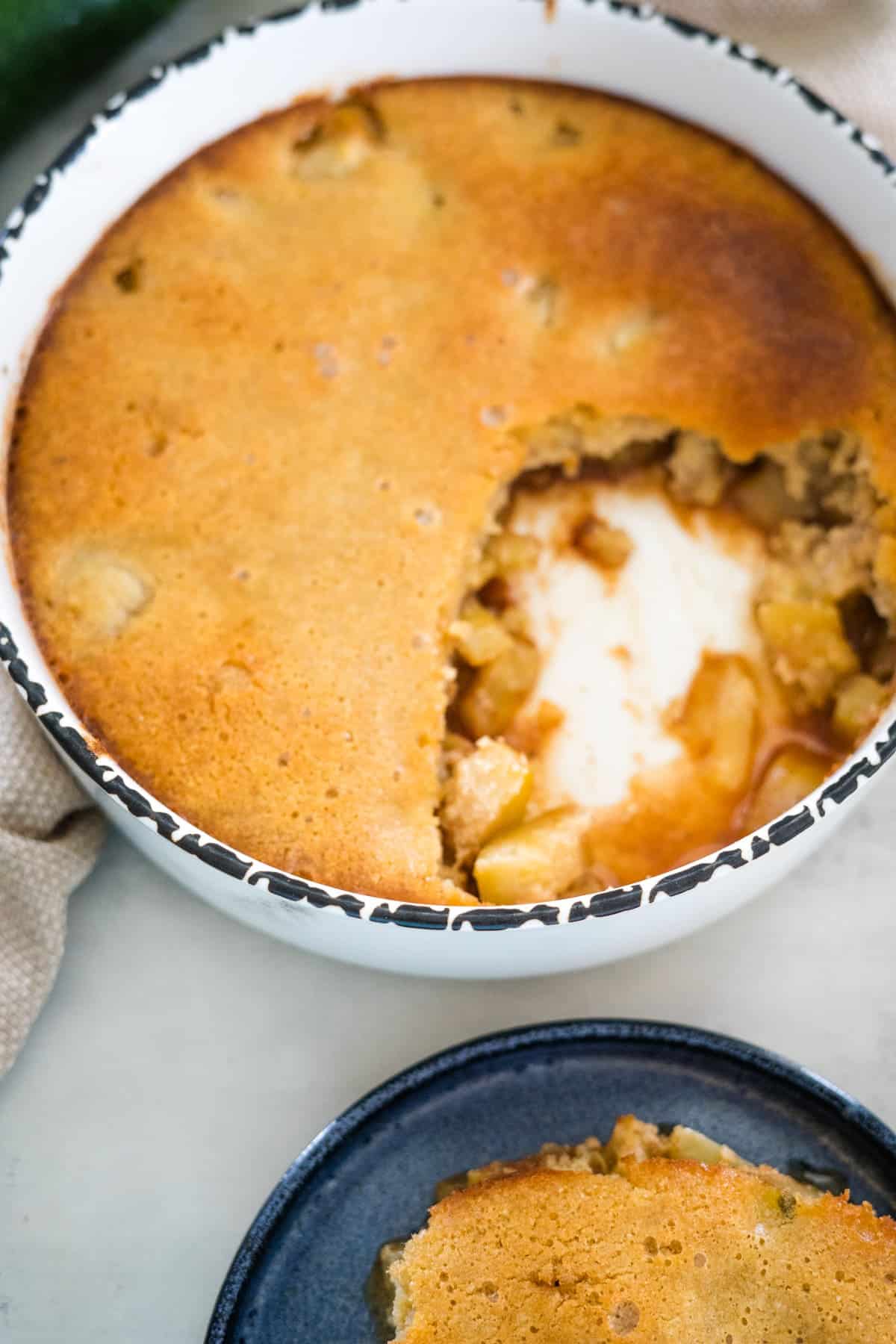 A baked apple cobbler in a white round dish with a portion scooped out, revealing the filling. A plate with a serving of the cobbler sits next to the dish.