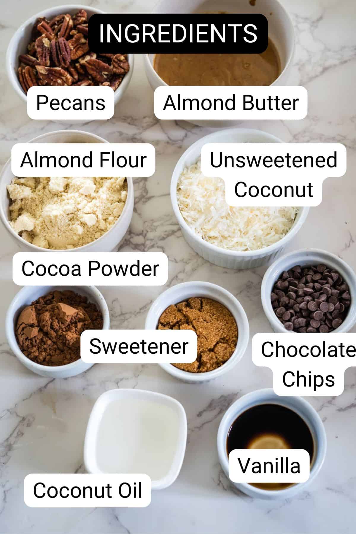 Various labeled ingredients including pecans, almond butter, almond flour, unsweetened coconut, cocoa powder, sweetener, chocolate chips, coconut oil, and vanilla, arranged on a marble surface.