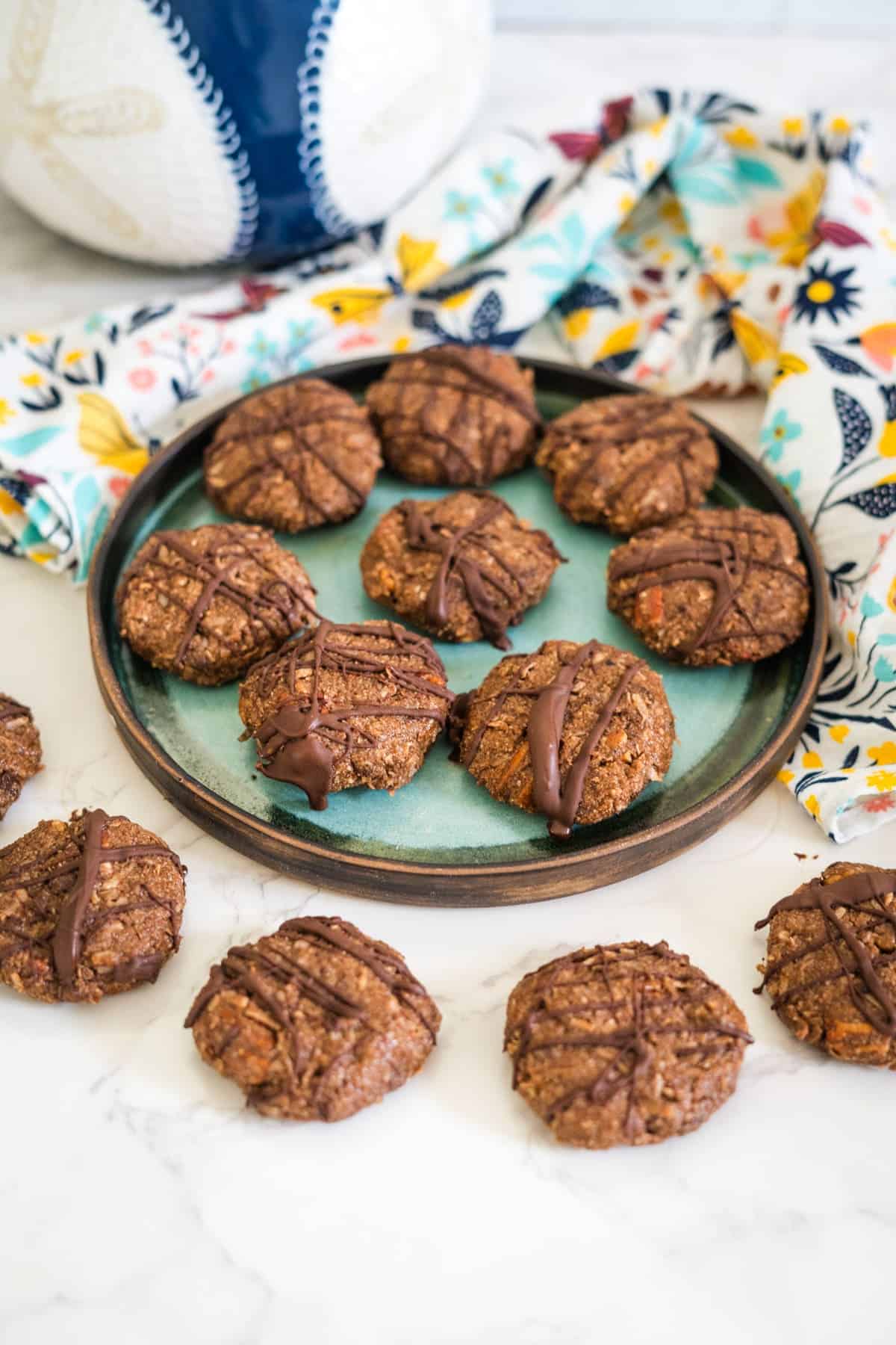 A plate with ten chocolate-drizzled, keto no bake cookies is on a marble surface, surrounded by a colorful floral cloth. Additional cookies are scattered around the plate.