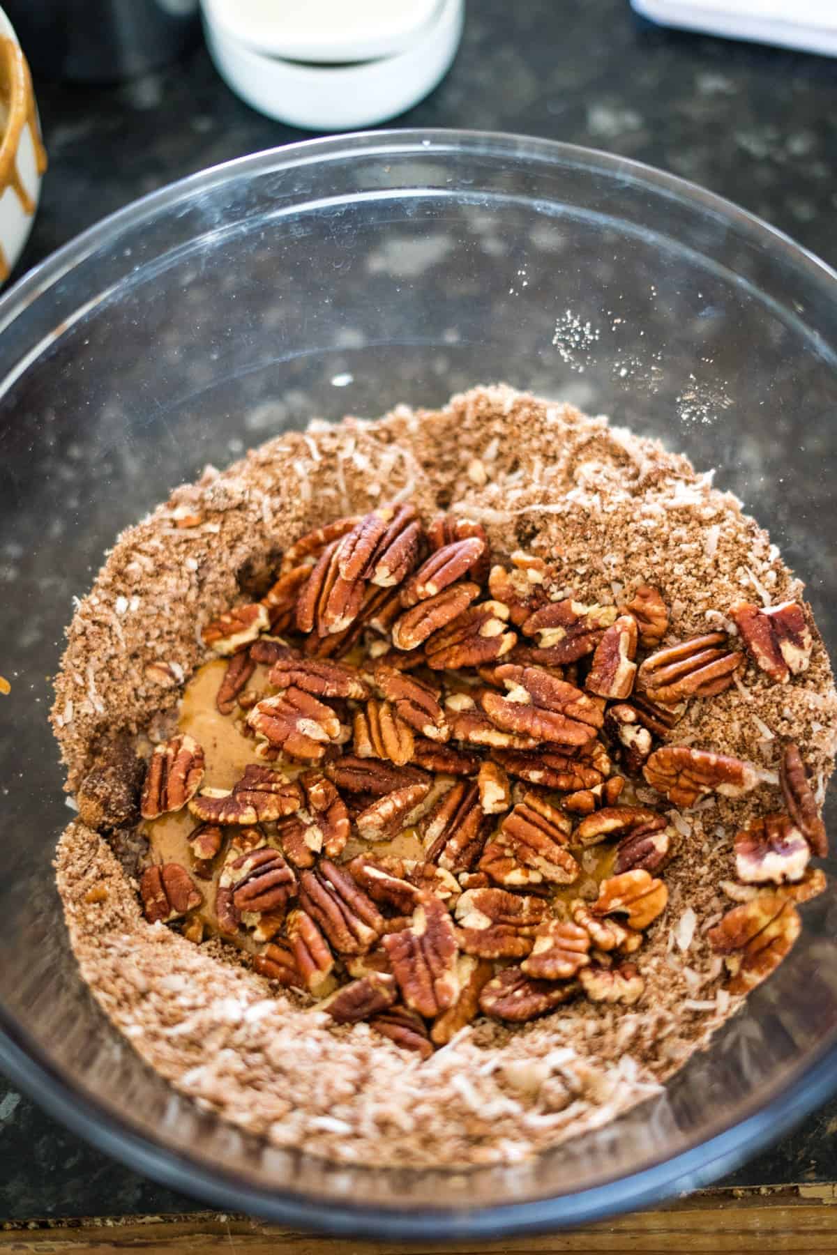 Glass bowl containing chopped pecans mixed with a blend of brown sugar and fine white granules, set on a dark countertop.