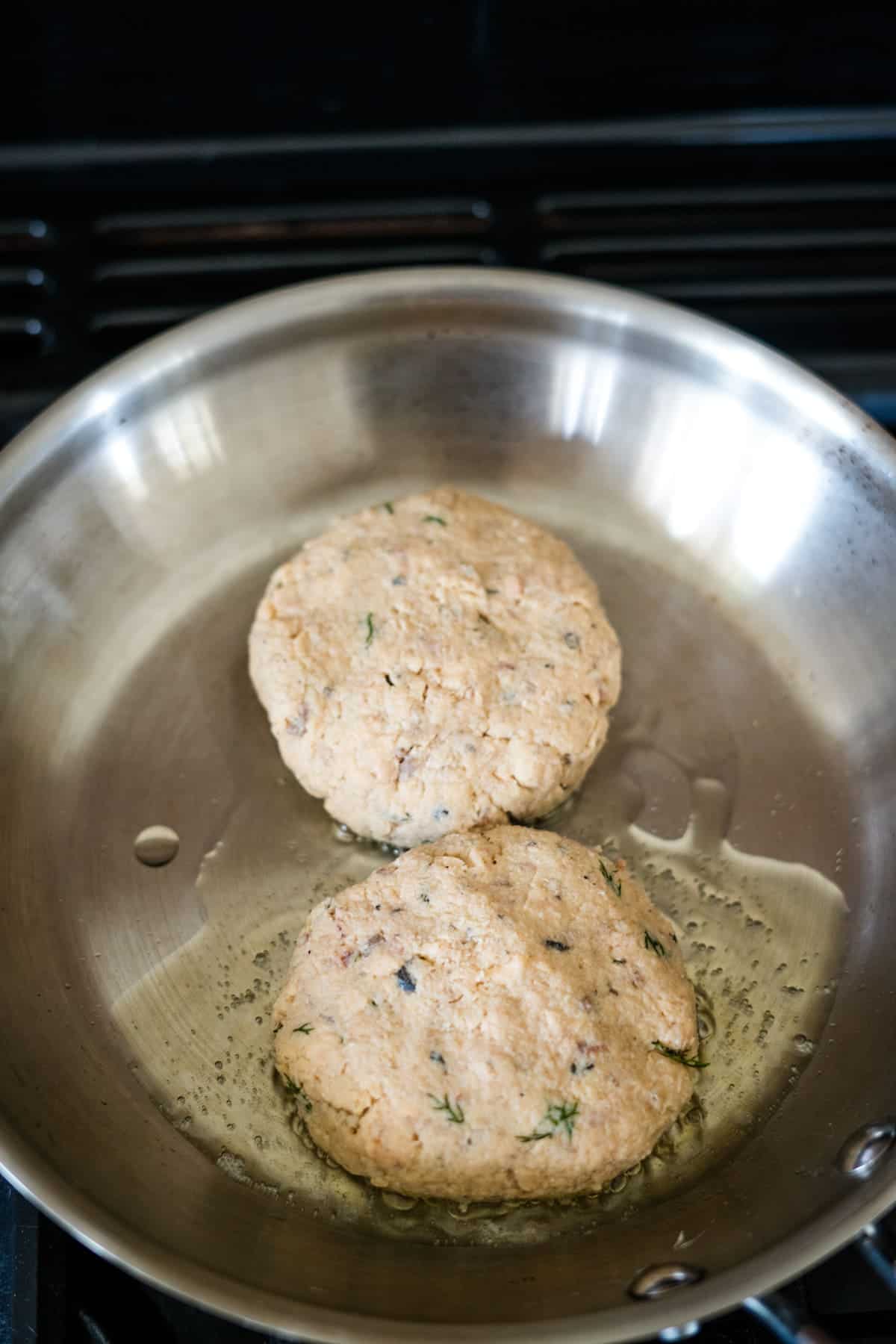 Two uncooked patties are frying in a stainless steel pan on a stovetop.