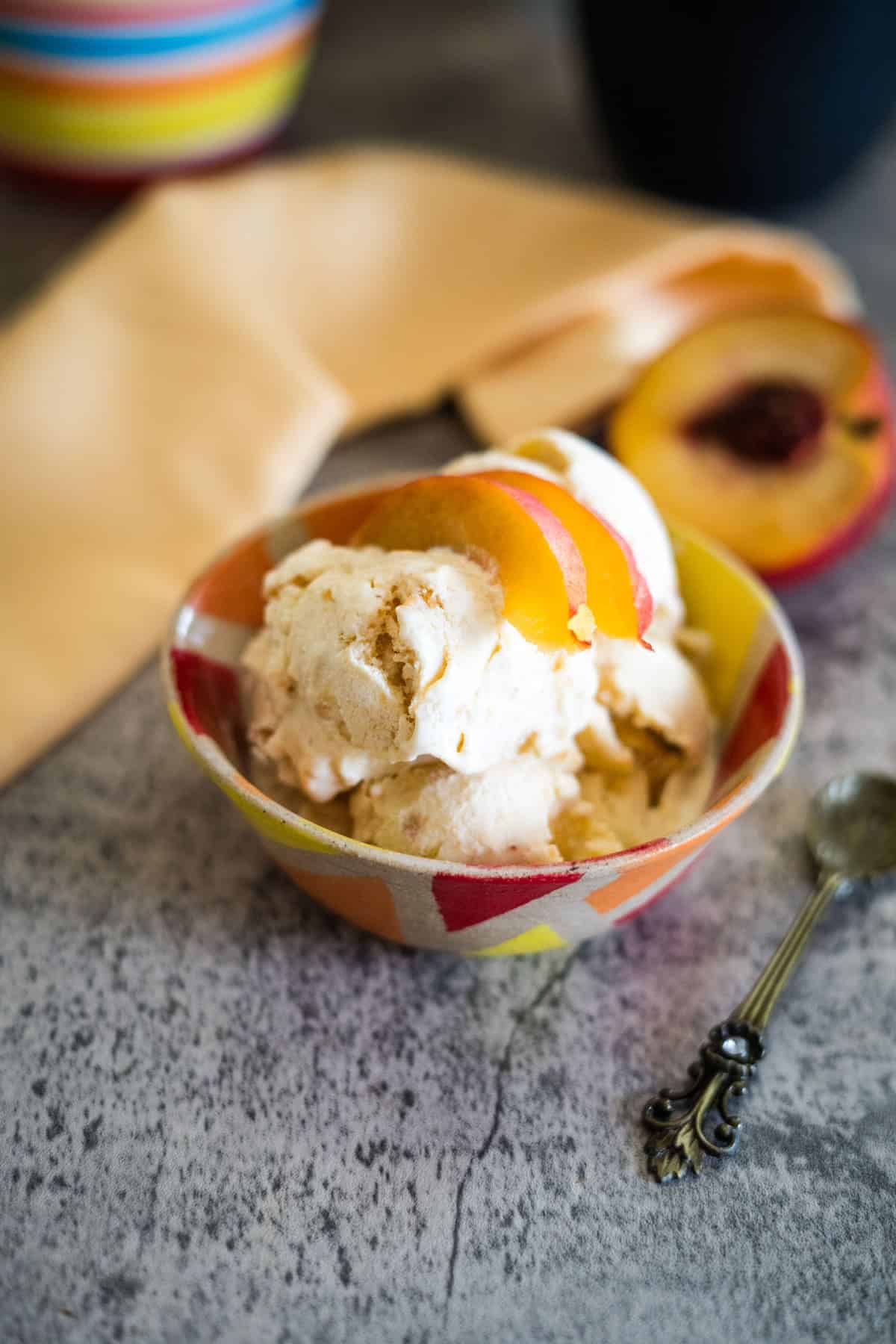 A bowl of peach ice cream with peach slices on top, placed next to a vintage spoon and a sliced peach on a gray surface.