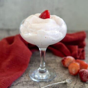 A dessert glass filled with strawberry mousse garnished with a strawberry slice, placed on a textured surface beside a red cloth and some whole strawberries.