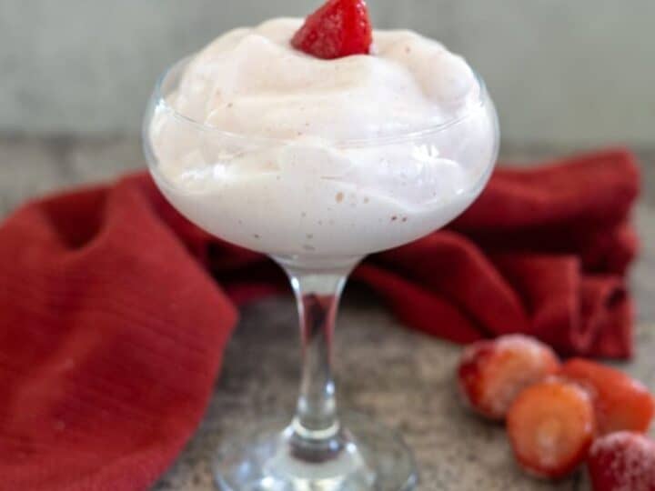 A dessert glass filled with strawberry mousse garnished with a strawberry slice, placed on a textured surface beside a red cloth and some whole strawberries.