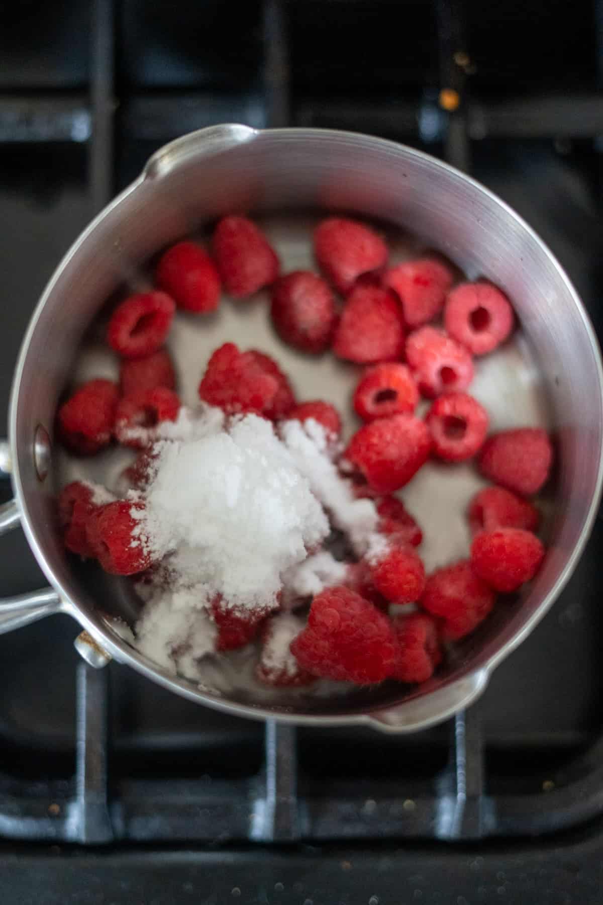 A metal saucepan on a stove contains raspberries and a pile of white granulated sugar.