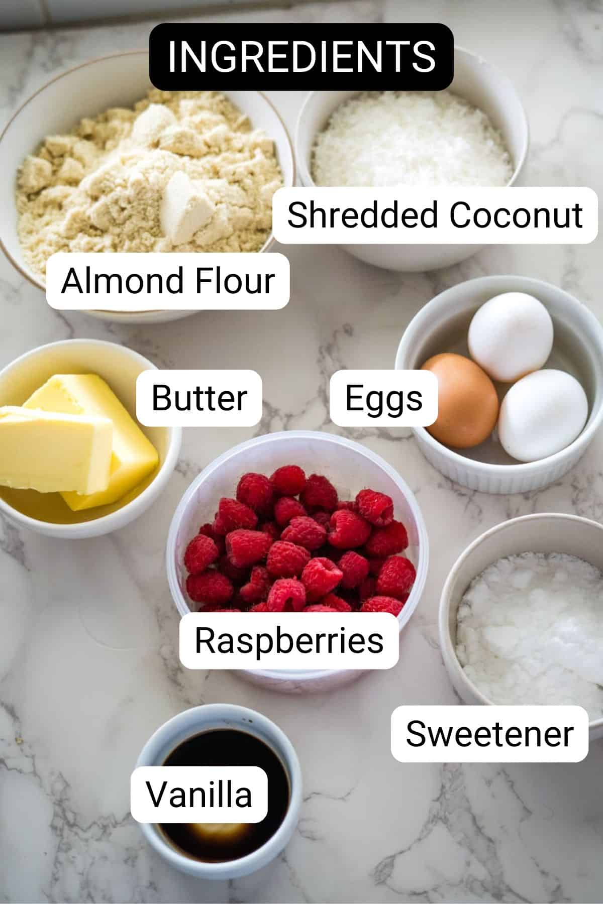 A variety of labeled ingredients on a marble countertop, including almond flour, butter, vanilla, shredded coconut, eggs, raspberries, and sweetener.