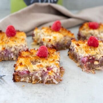 Close-up of four coconut raspberry bars topped with fresh raspberries, arranged on a white surface with a beige cloth in the background.