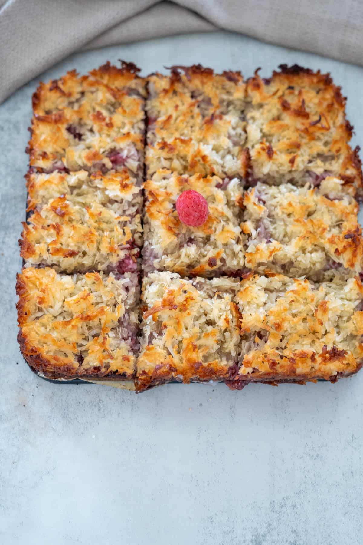 Square pieces of baked coconut dessert topped with a single raspberry on a light surface.