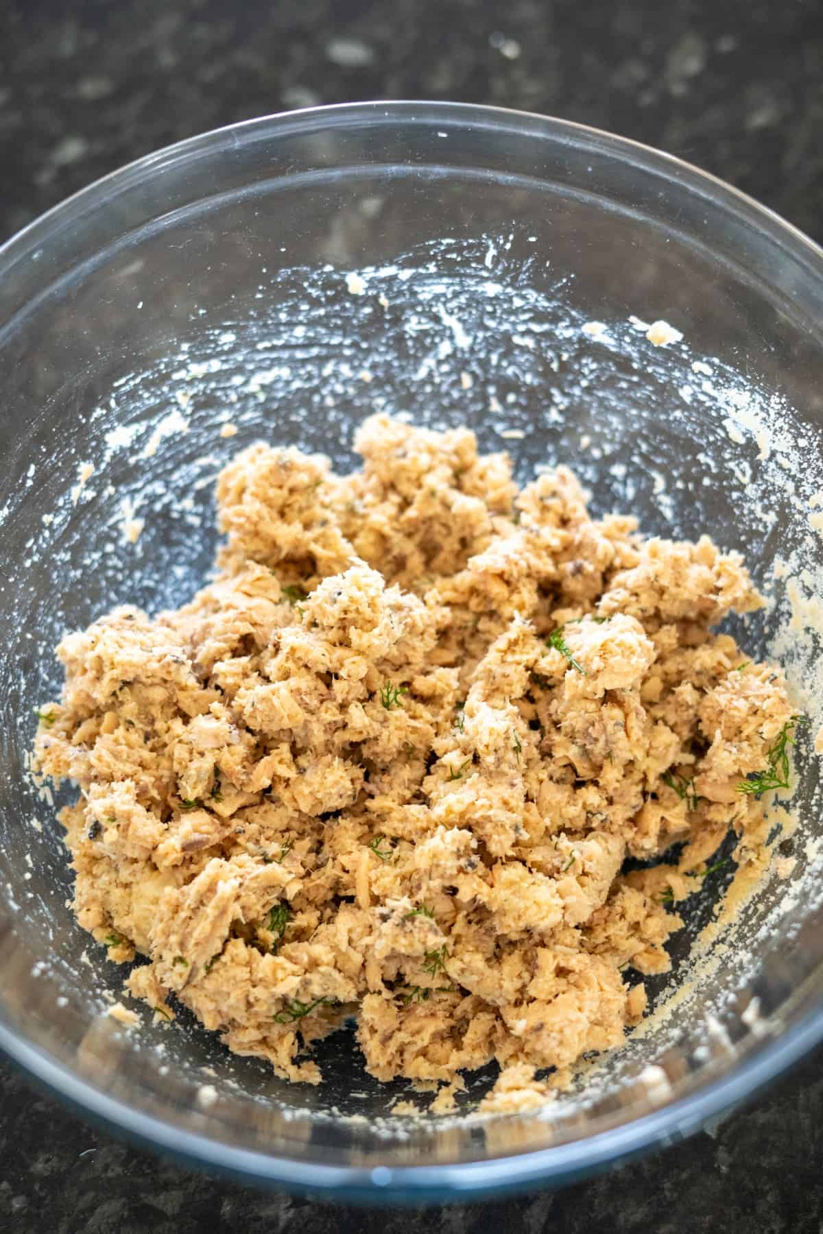 A large clear mixing bowl filled with a mixture of ingredients, resembling a dough or stuffing, on a dark, speckled countertop.