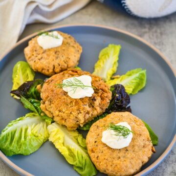 A plate holds three crab cakes garnished with cream and dill, served on a bed of mixed lettuce leaves.