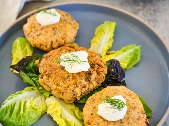 A plate holds three crab cakes garnished with cream and dill, served on a bed of mixed lettuce leaves.