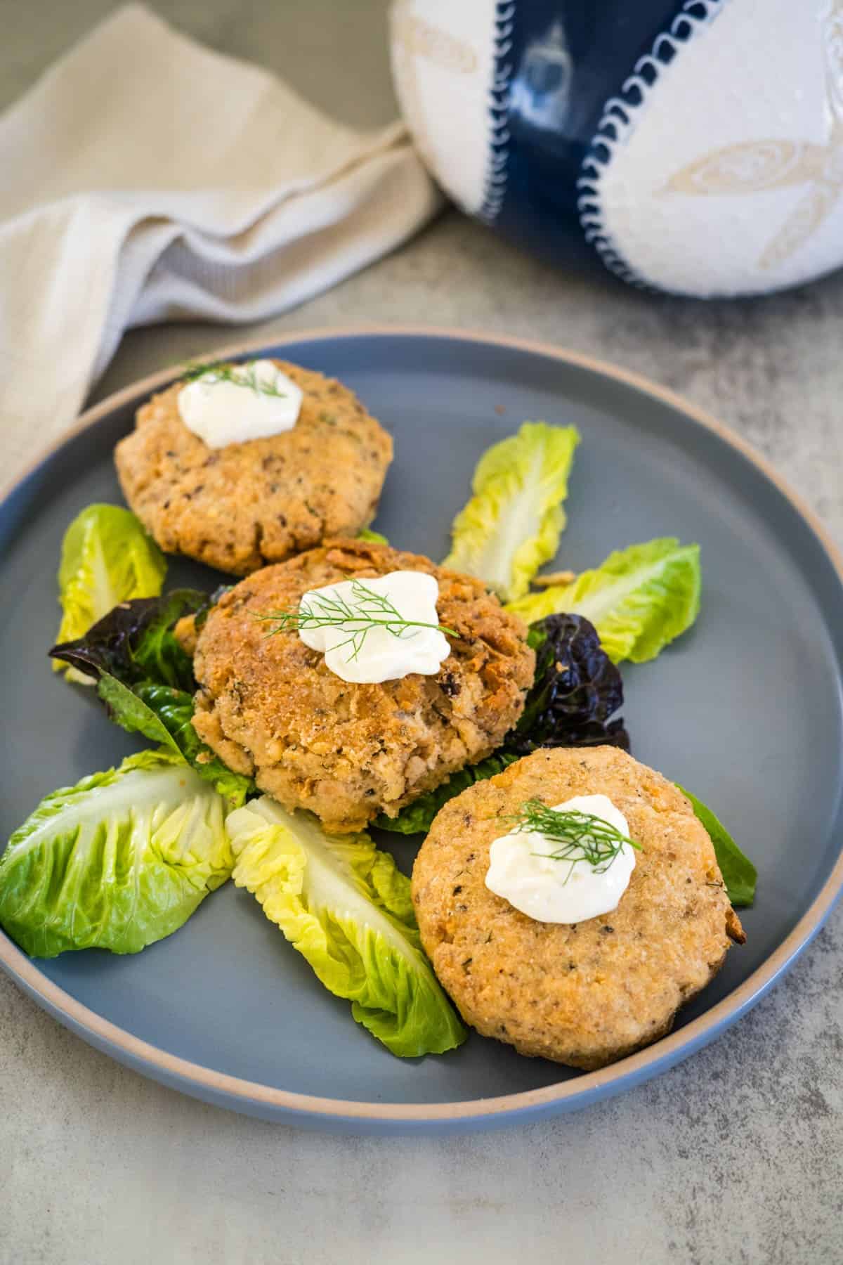 Three salmon cakes topped with dollops of sour cream are served on a bed of mixed lettuce on a gray plate, with a ceramic vase and a light-colored napkin in the background.