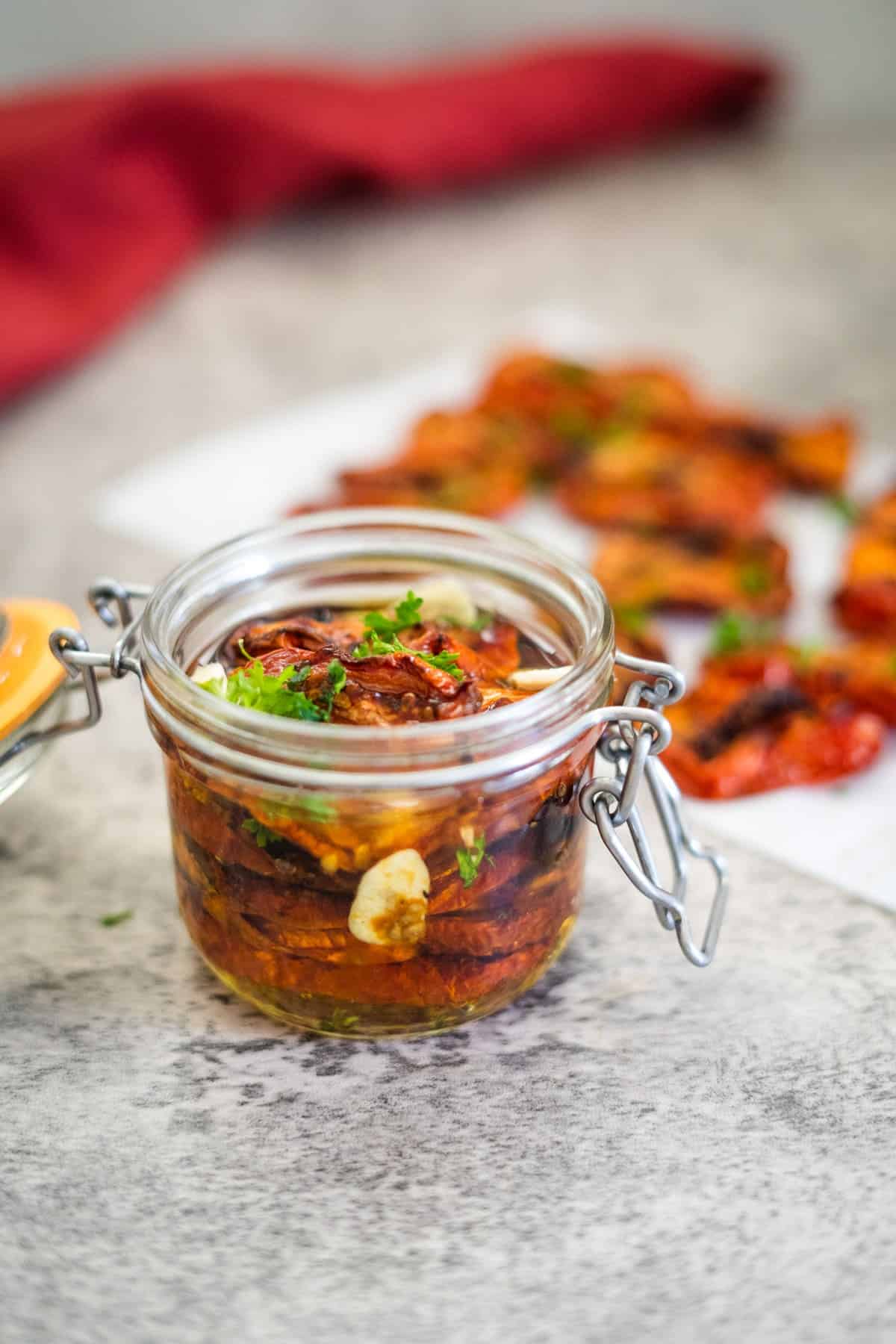 A glass jar filled with sun-dried tomatoes, garnished with herbs and garlic slices, sits on a countertop. A red cloth is visible in the background.