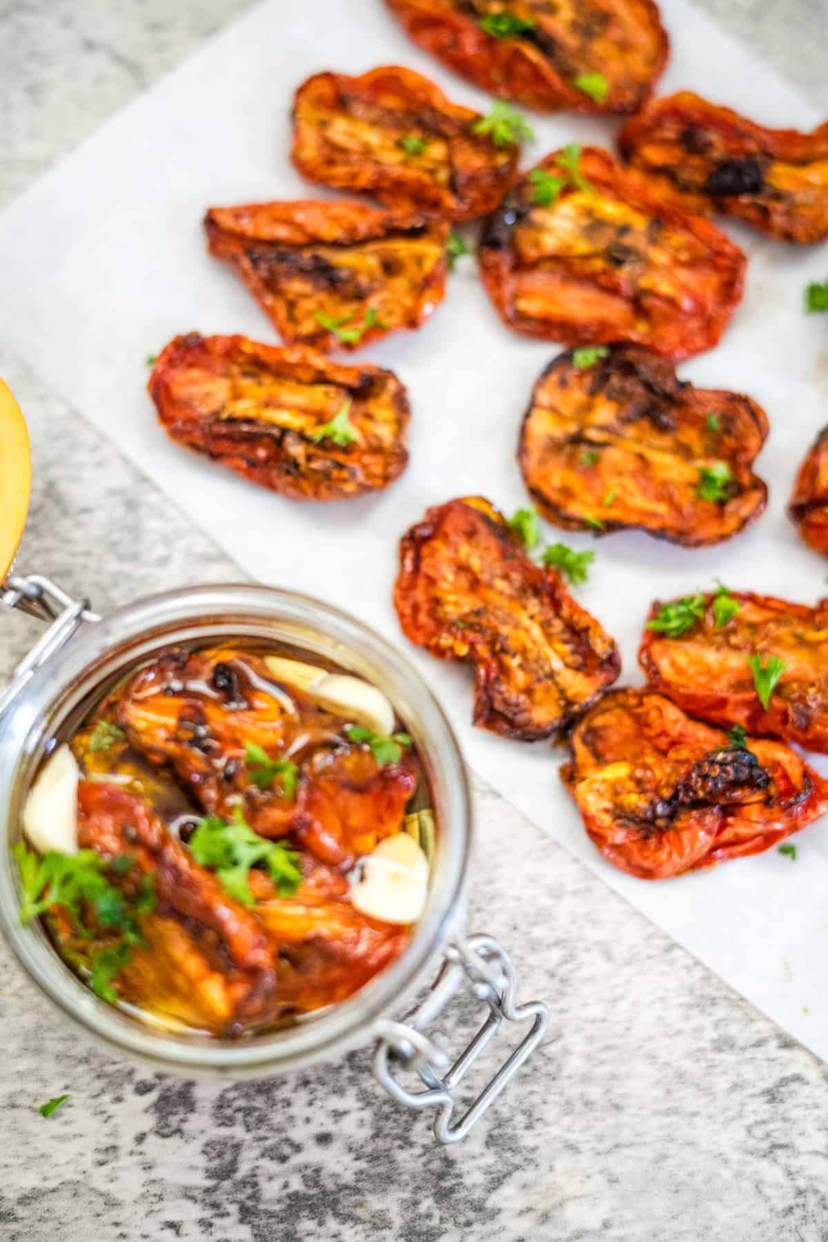 An overhead view of air fryer sun-dried tomatoes on parchment paper, with a glass jar filled with more sun-dried tomatoes, garlic, and herbs.