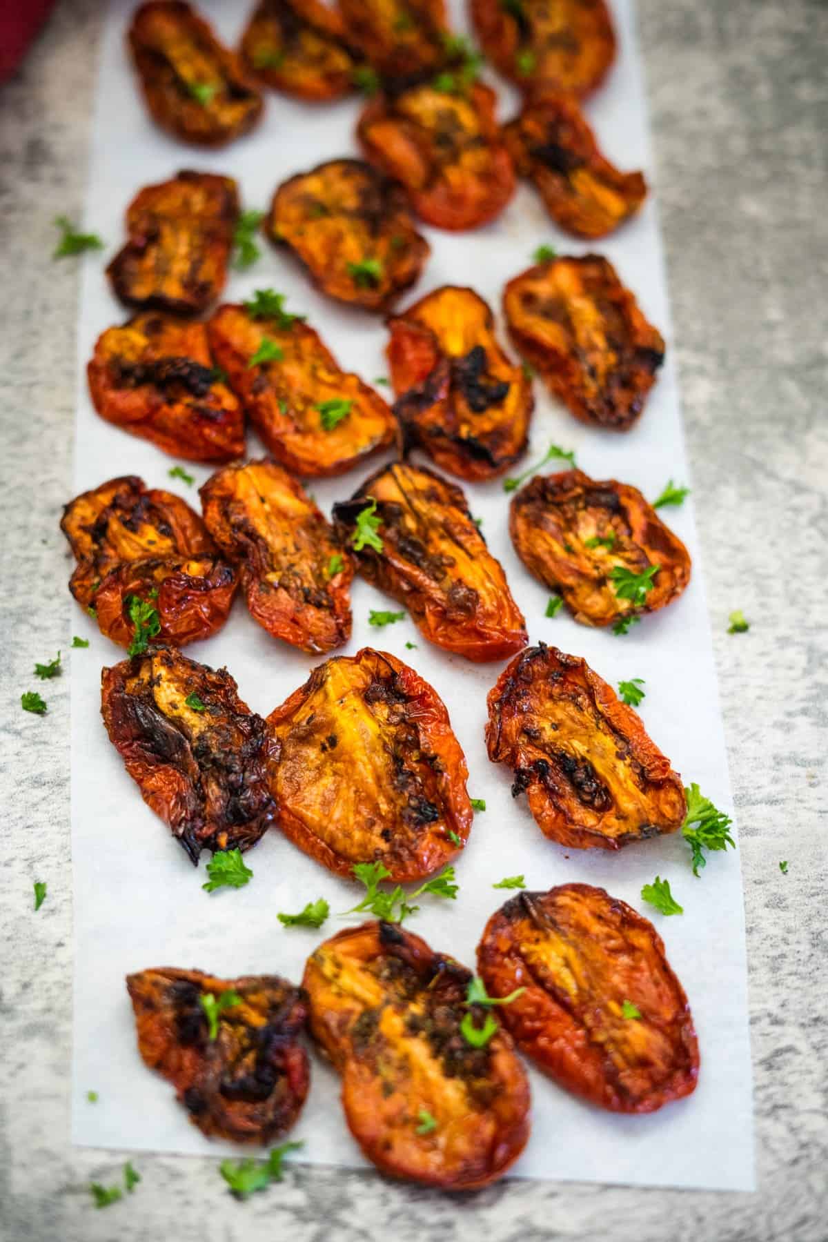 A parchment paper with roughly twenty pieces of air fryer sun dried tomatoes garnished with chopped parsley, arranged in a scattered pattern on a light grey surface.