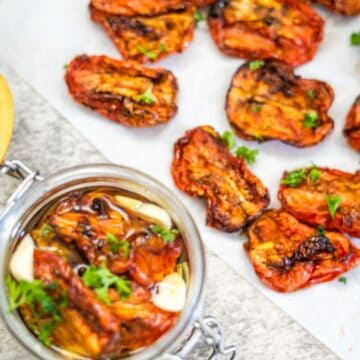 Several sun-dried tomato halves on parchment paper, garnished with herbs, with a jar of sun-dried tomatoes in oil and garlic on the side.