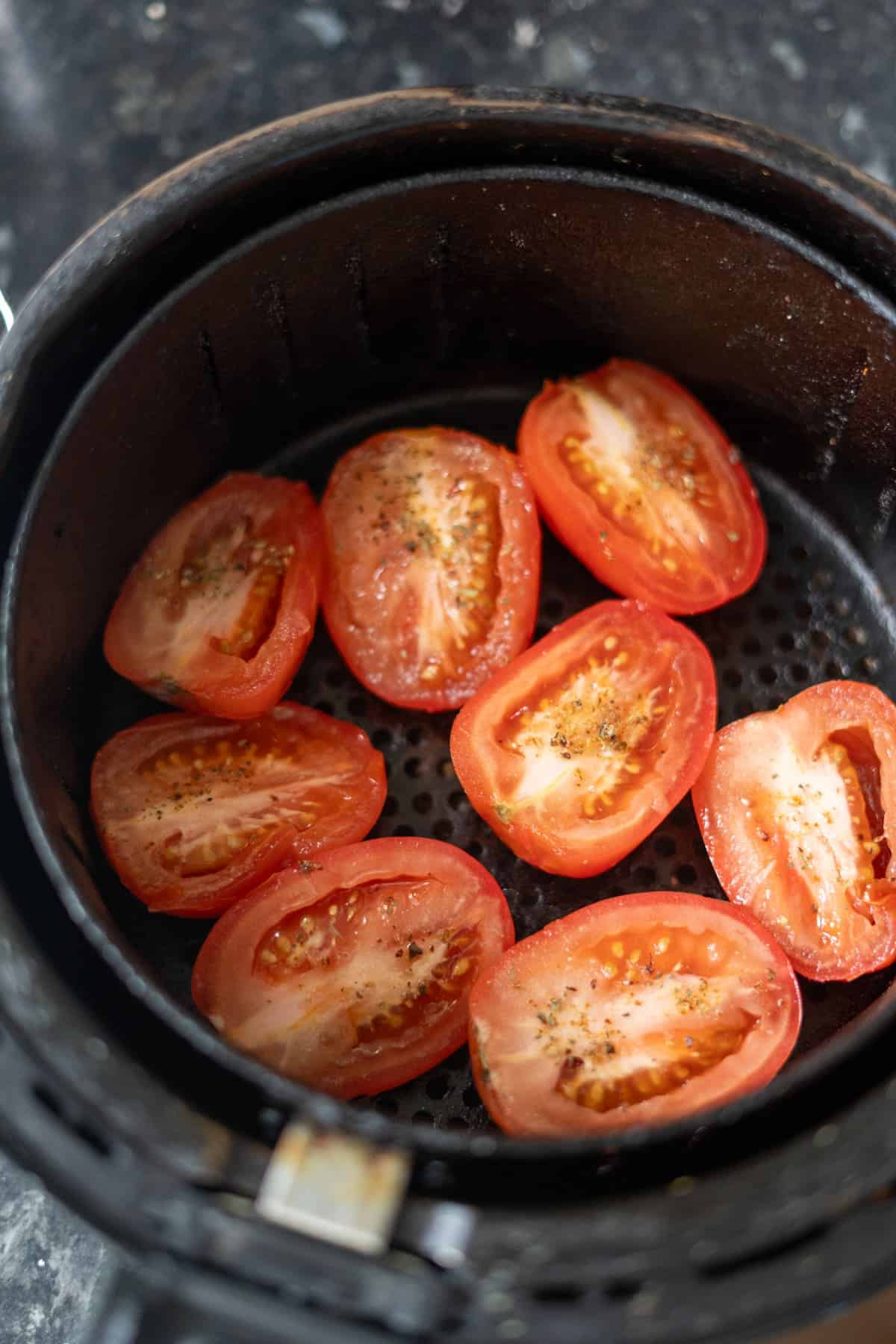 Halved tomatoes seasoned with salt and pepper placed in the basket of an air fryer, creating delicious air fryer sun-dried tomatoes.