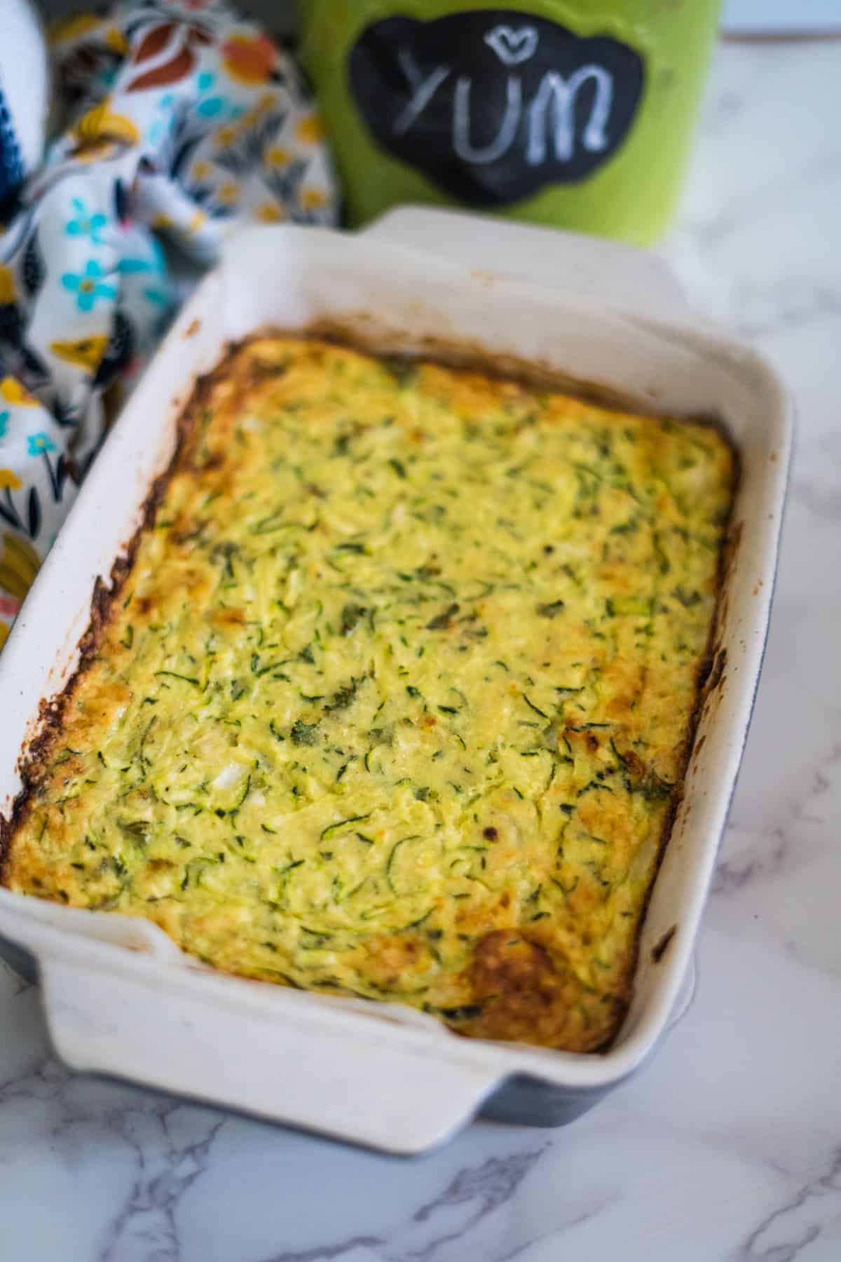 A baked casserole dish filled with a golden-brown keto zucchini slice is placed on a marble countertop. A chalkboard label in the background reads "YUM".