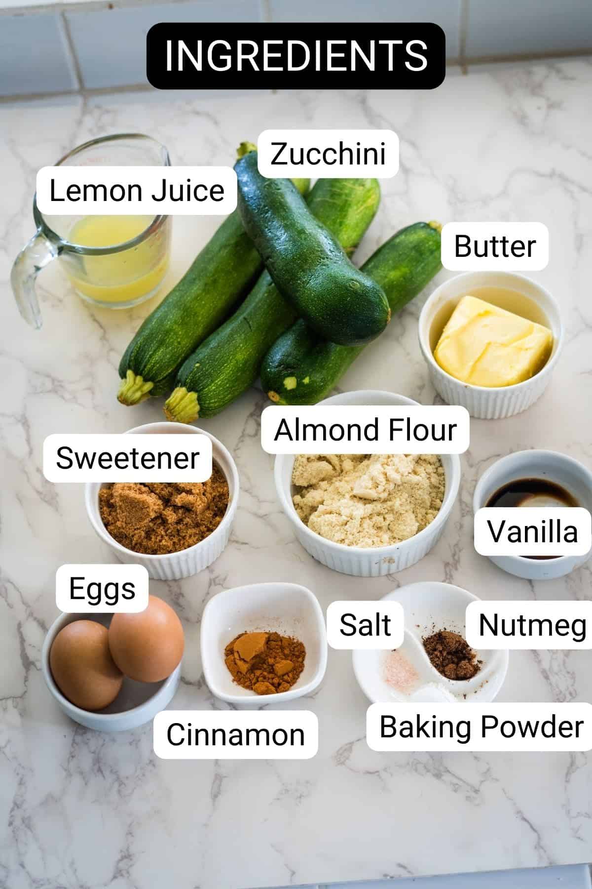 Ingredients for a zucchini recipe are arranged on a marble countertop: zucchini, butter, lemon juice, almond flour, sweetener, vanilla, eggs, salt, nutmeg, baking powder, and cinnamon.