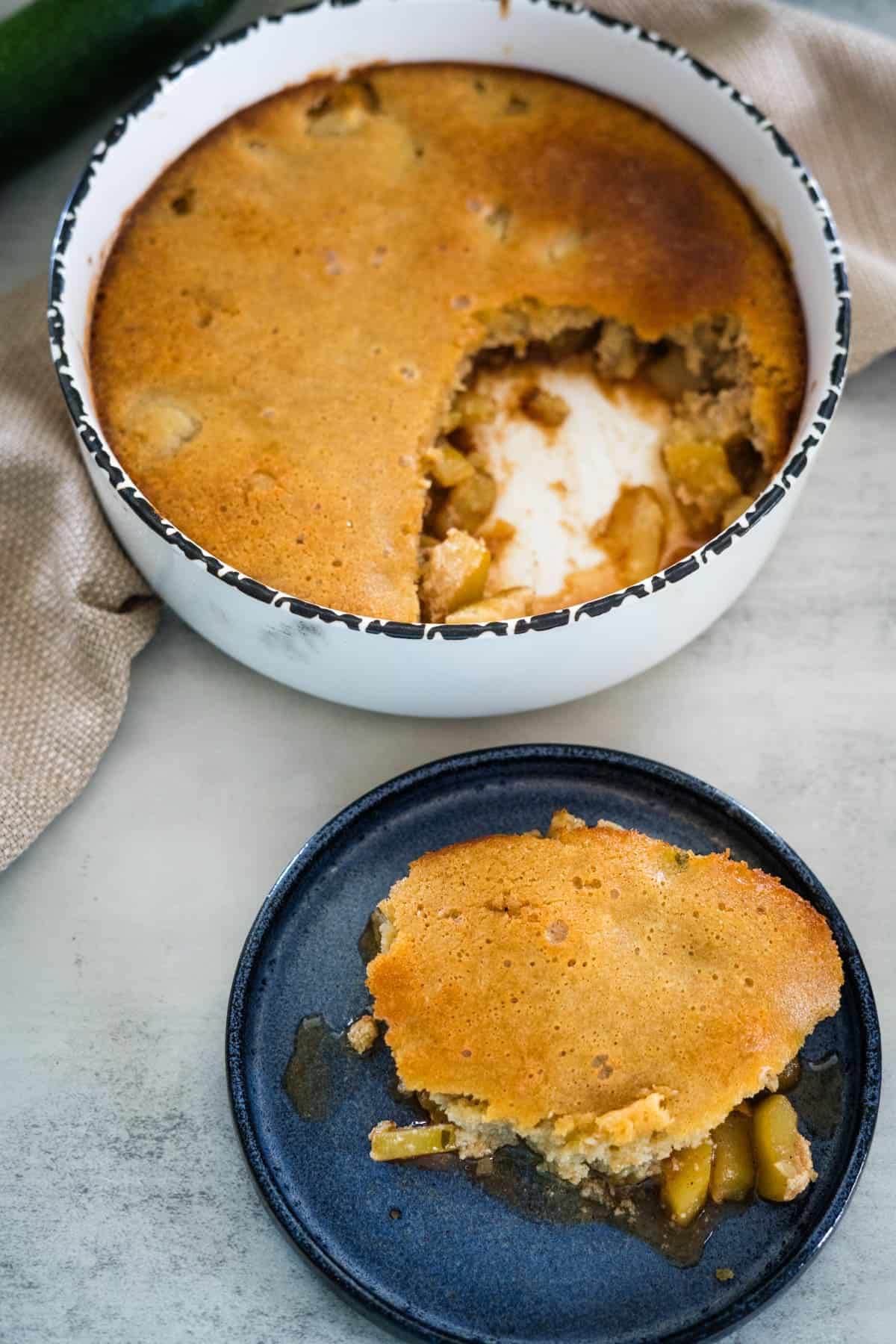 A round baking dish filled with a partially eaten golden brown cobbler and a separate serving on a dark plate, showing fruit filling inside.